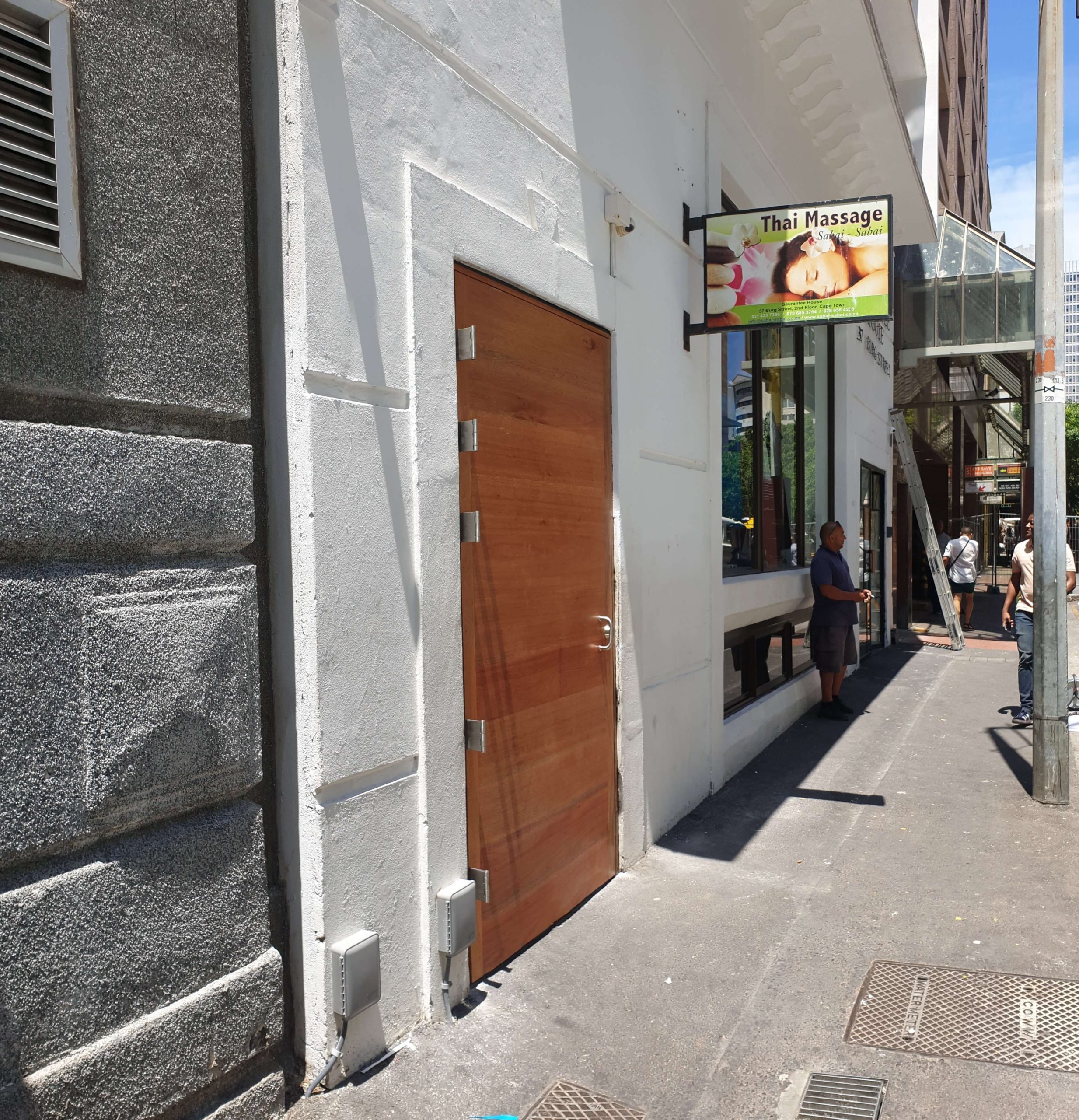 Sidewalk view of a building with a Thai massage sign and a boarded-up door. Several people on the street, including a man in a dark shirt and shorts, and a ladder leaning against the building.