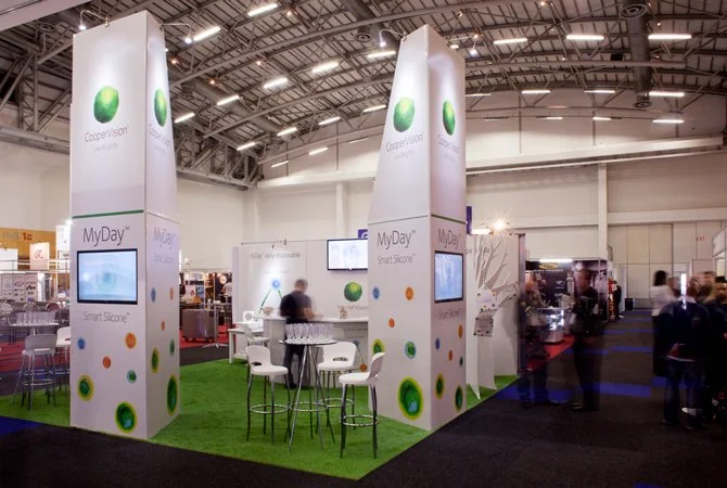 Trade show booth with white panels featuring green and orange circular patterns, a green turf floor, tables and chairs, and screens displaying company branding for Coppevision and MyDay.