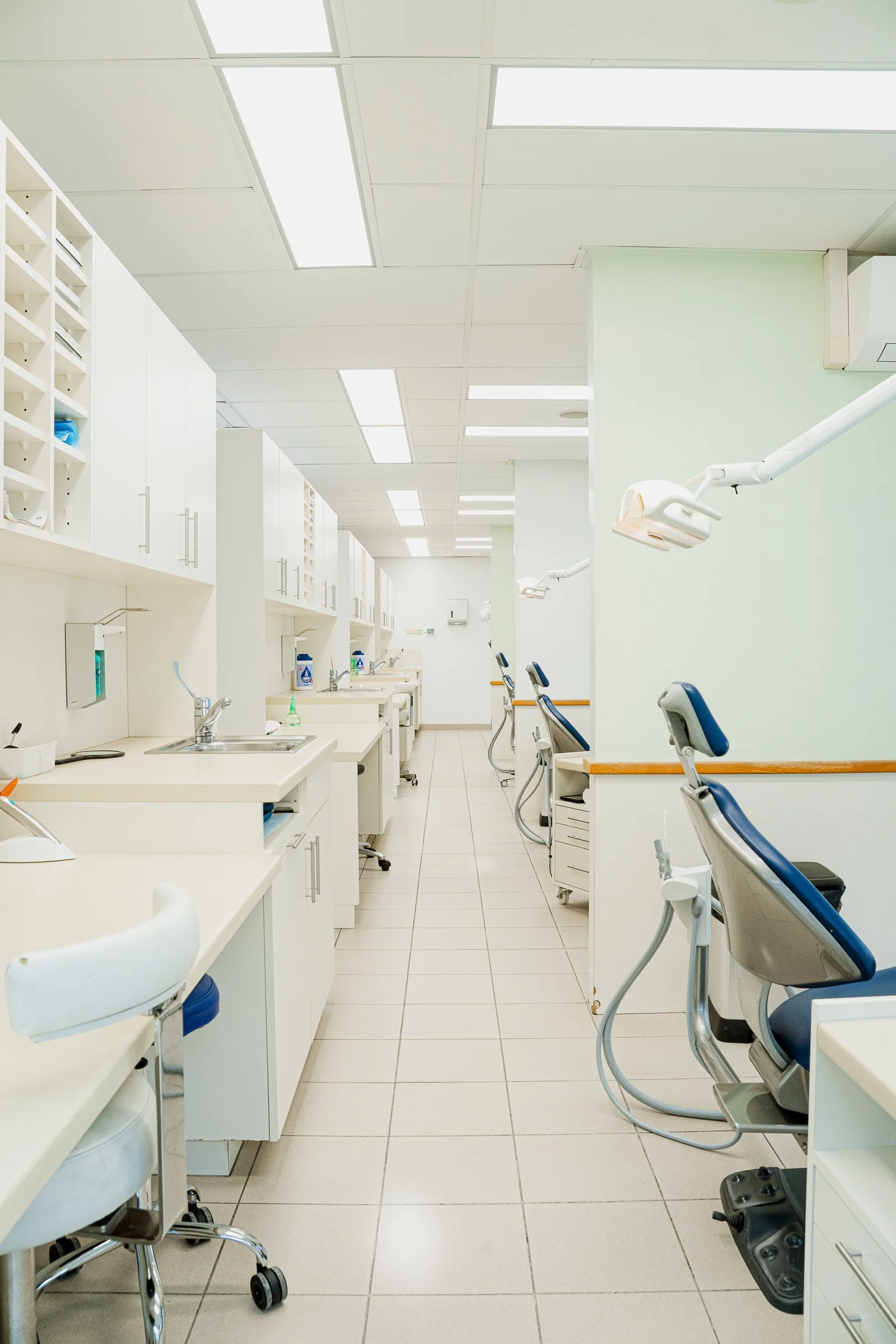 A dental clinic operatory room with multiple dental chairs, overhead lights, sinks, and cabinets.