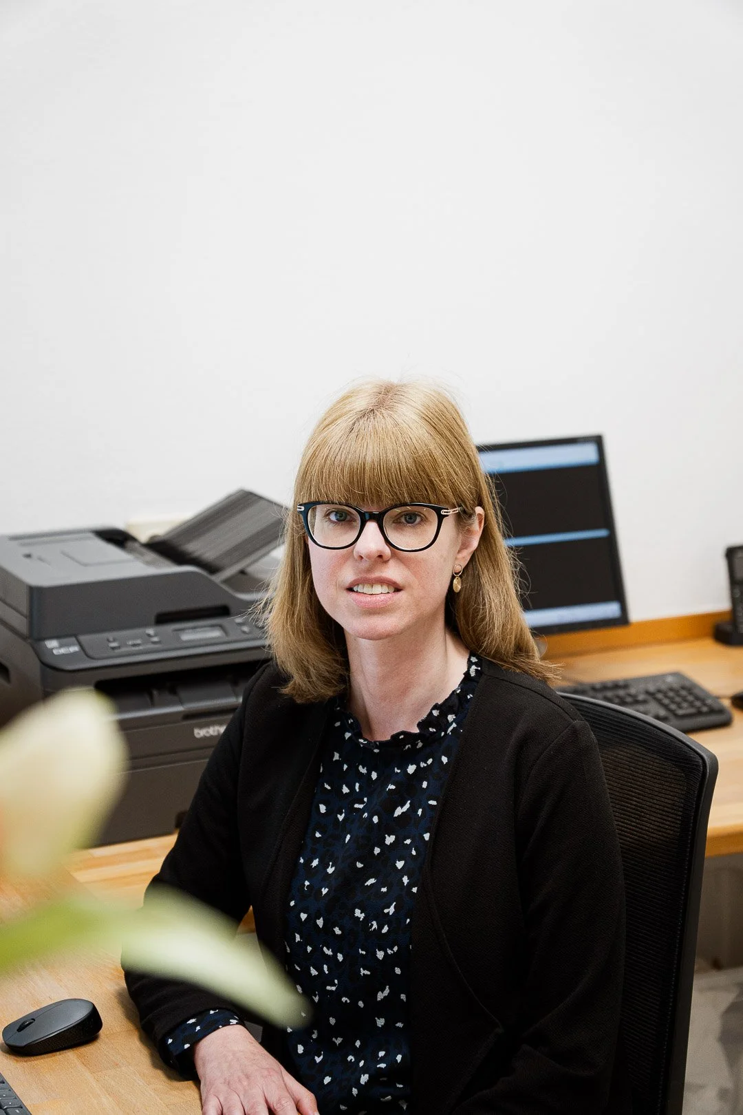 A woman with shoulder-length blonde hair and glasses, wearing a black blazer and a navy blouse with a small white pattern, sitting at a desk in an office environment. There is a printer, a computer monitor, a keyboard, a mouse, and a black phone on the desk.