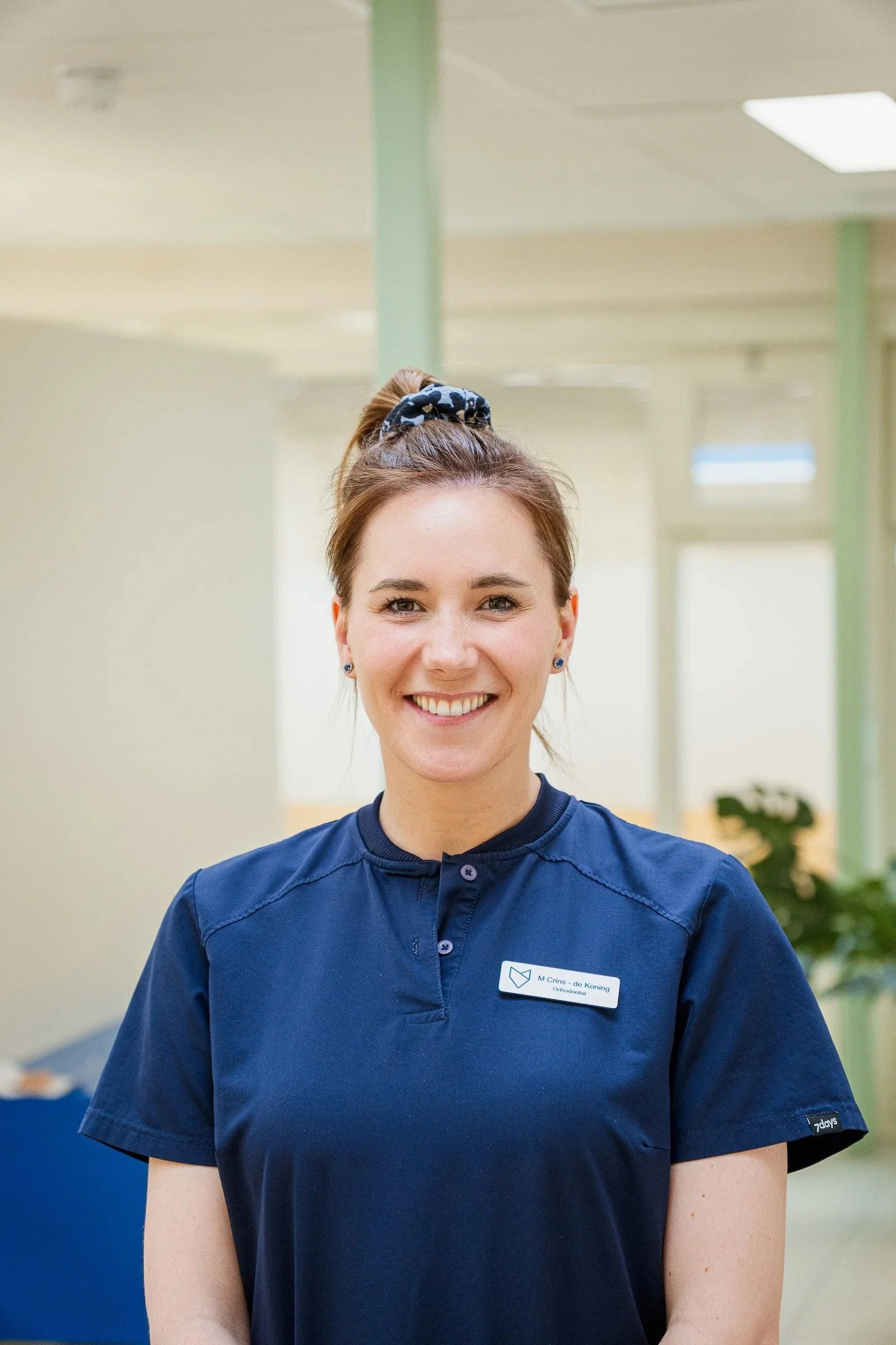 Smile of a young woman with freckles wearing a navy scrub top with a name tag in a clinical or office setting.