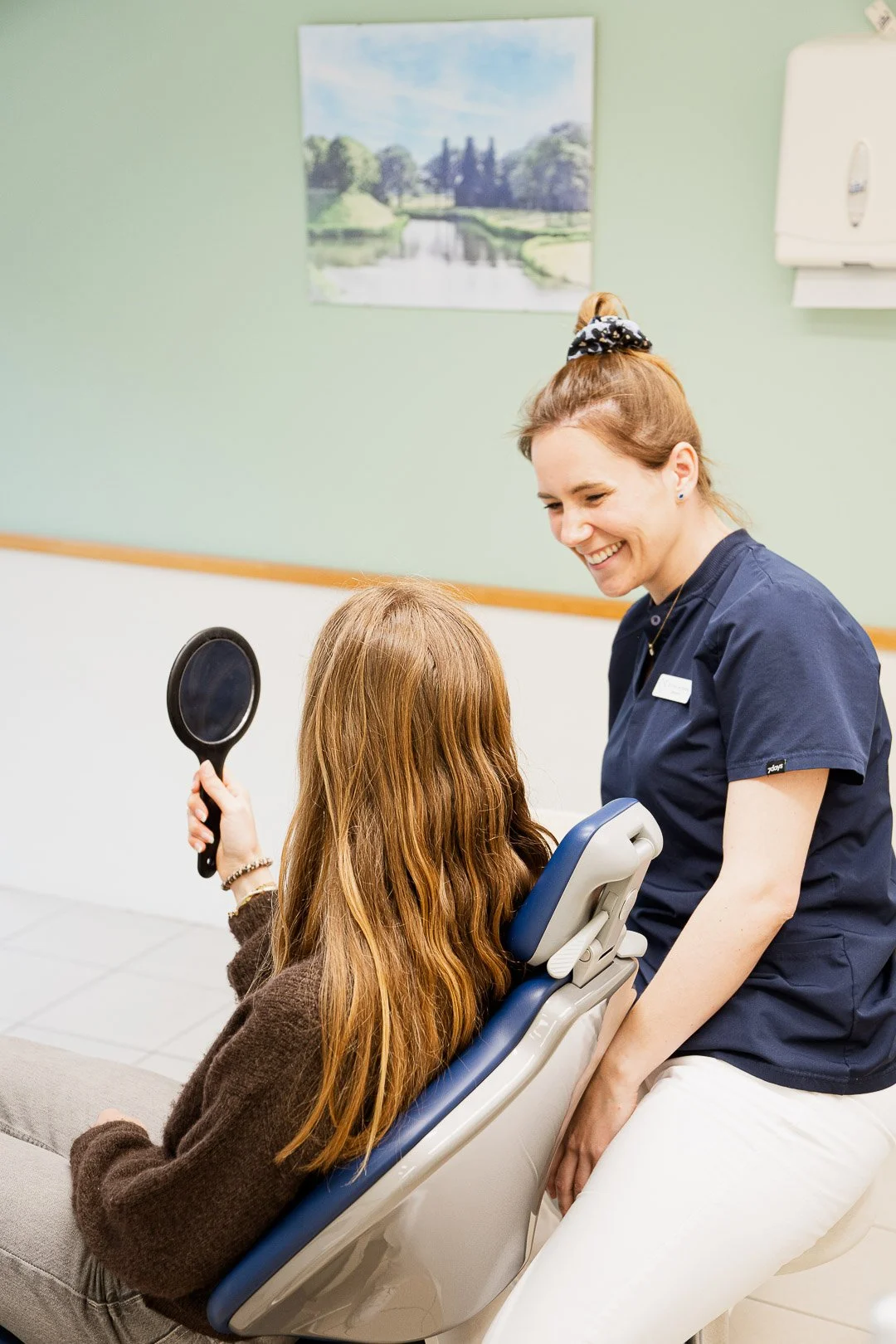 A young girl with long red hair sitting in a dental chair, holding a mirror, while a smiling dental hygienist in navy scrubs leans over her.