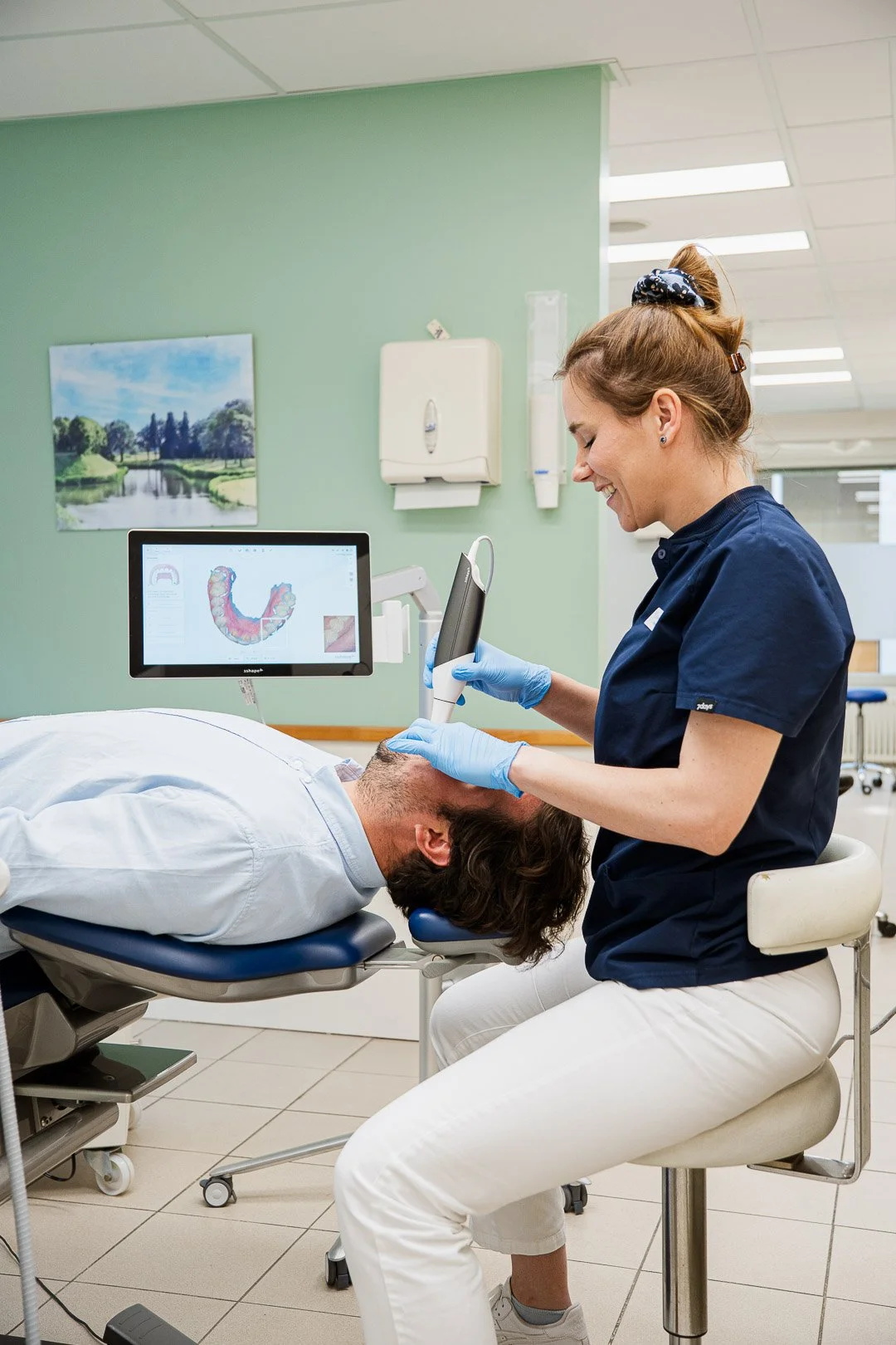 A dentist using a scanning device on a patient who is lying back in a dental chair, with a monitor showing a digital image of teeth.