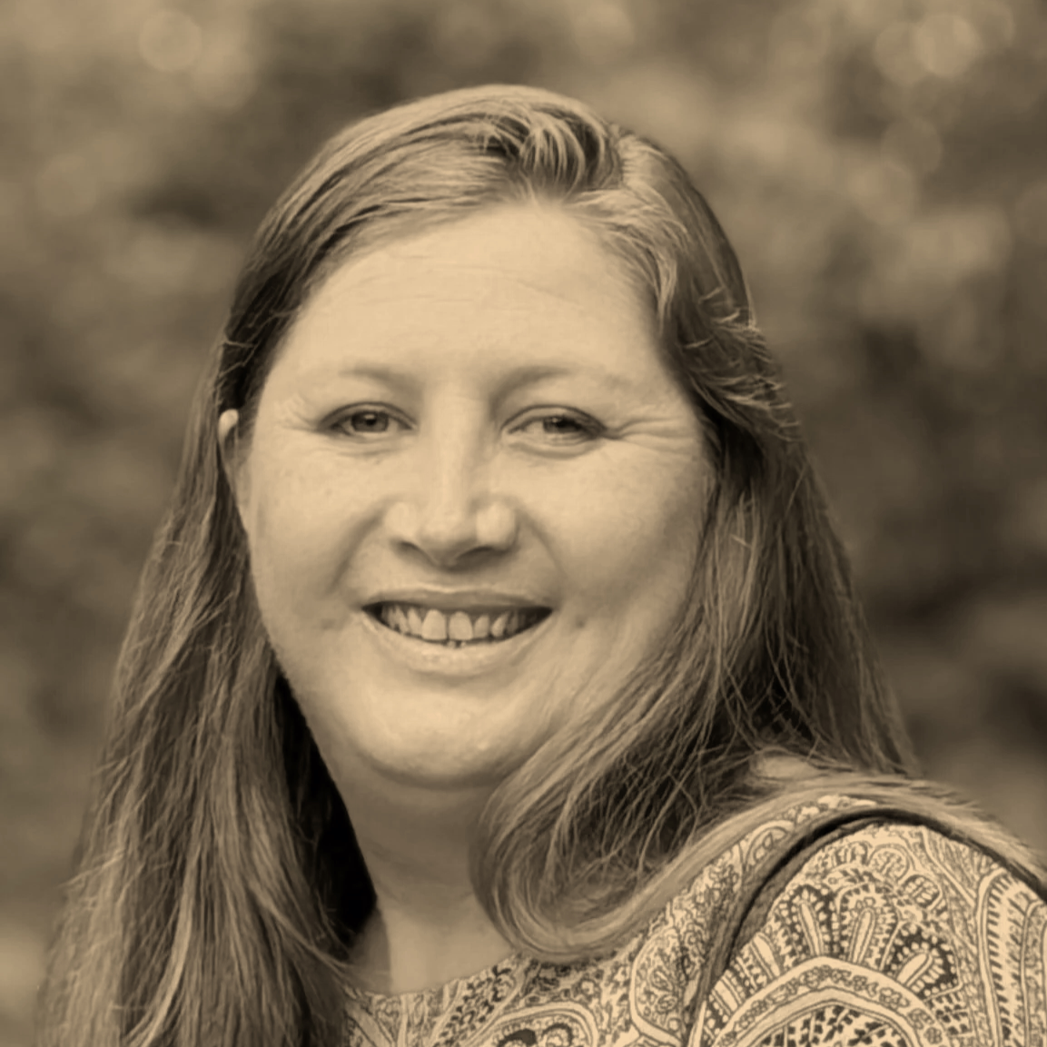 A black and white headshot of Nikki smiling, wearing a paisley shirt, against a natural backdrop.