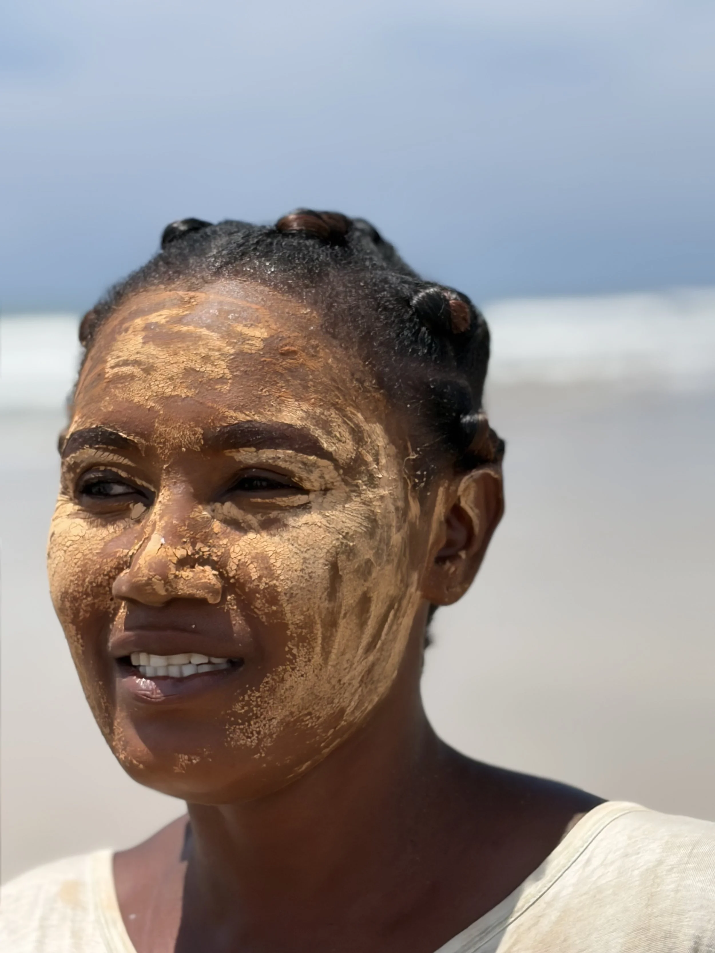 An portrait of an African woman with a mud protection layer on her face looks away from the camera towards the horizon. Her expression is a soft countenance and warm smile. The background is blurred, beach sand meets ocean which in turn meets sky.