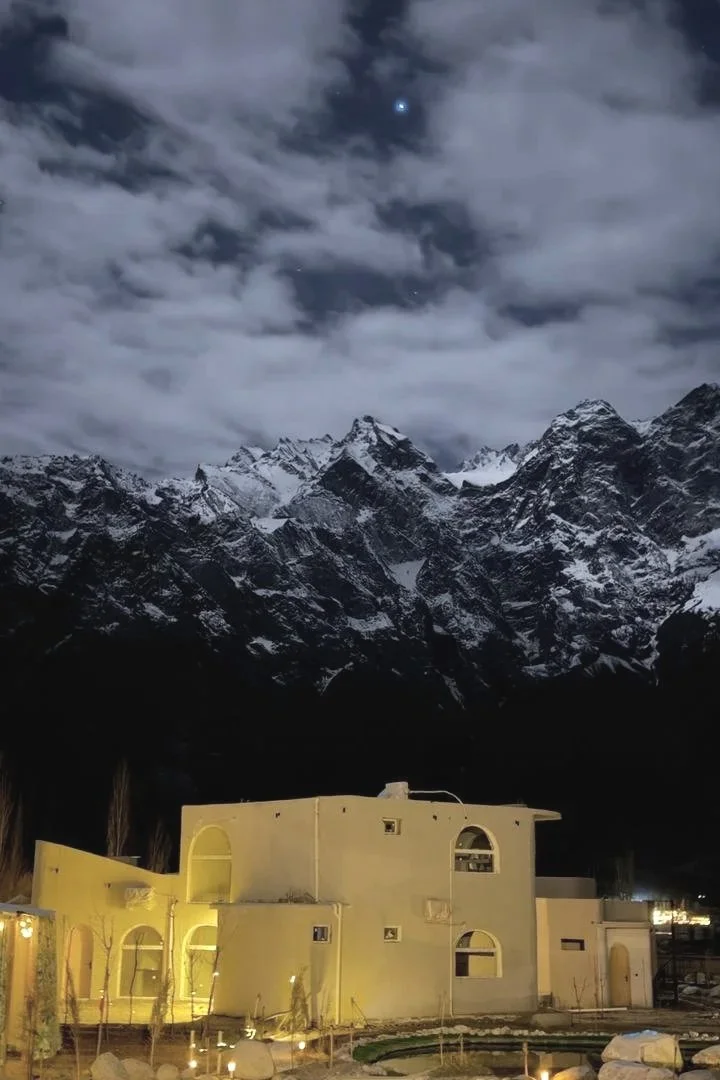 Nighttime view of a modern white house with rounded windows, set against snow-capped mountains under cloudy sky.