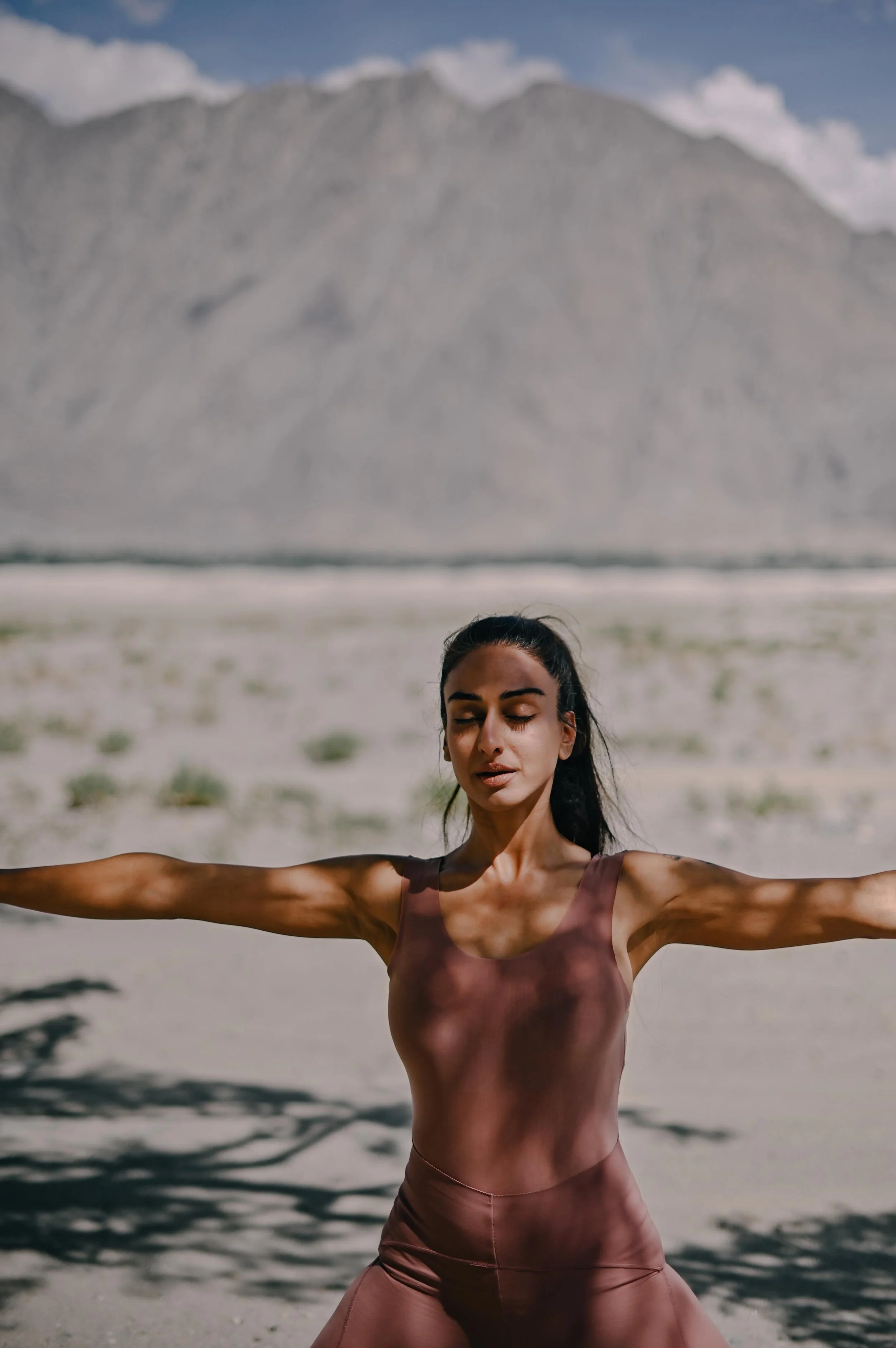 A woman standing on a beach with her arms outstretched, wearing a brown sports bra and pants, with mountains in the background and clouds in the sky.