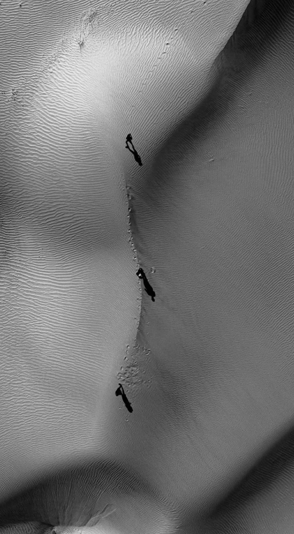 Aerial view of sand dunes with wind patterns, featuring three small figures walking along a ridge, creating shadows on the dunes.