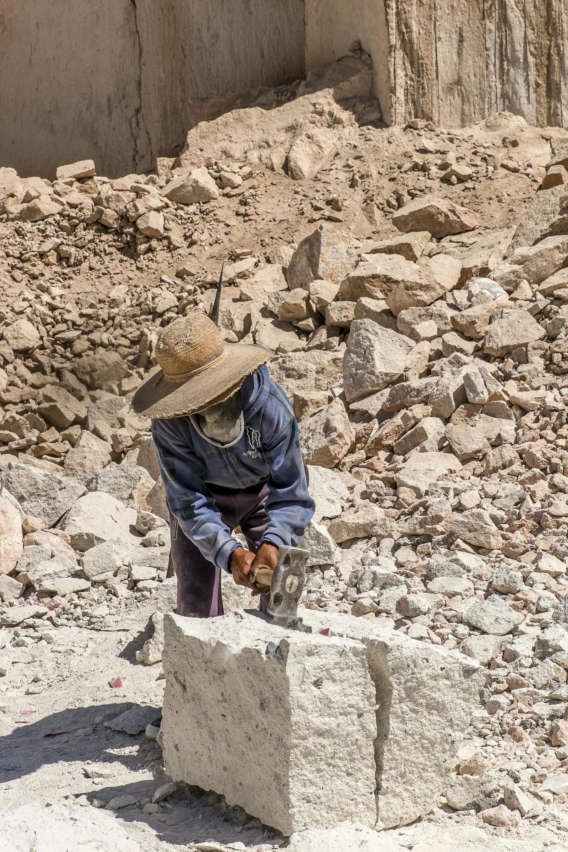 A worker wearing a wide-brimmed straw hat, long sleeves, and face covering, using a pickaxe to break large rocks in a sunny, rocky outdoor environment, possibly a quarry or excavation site.