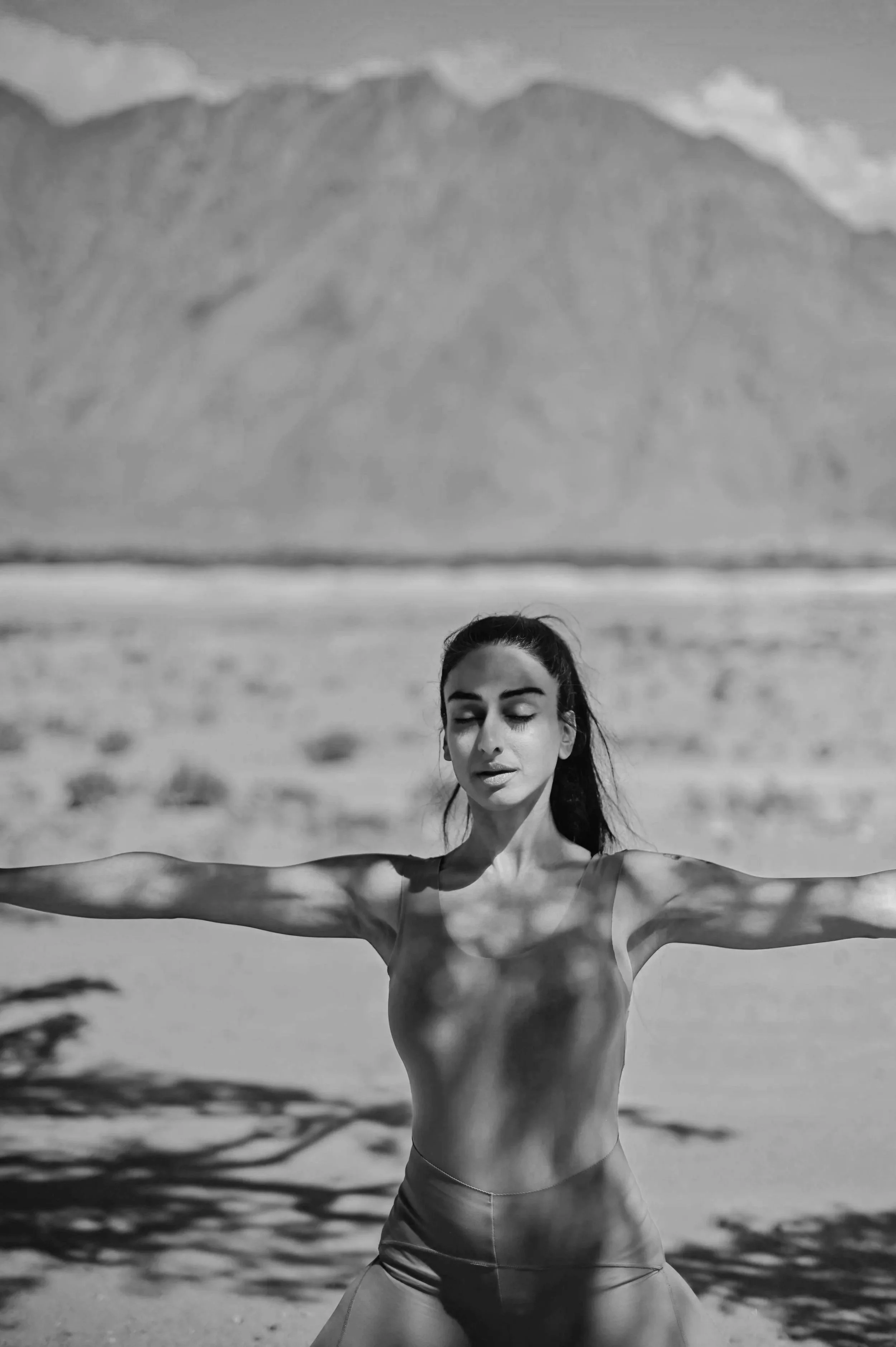 A woman practicing yoga on the beach with her arms extended outward, eyes closed, and a mountain range in the background.