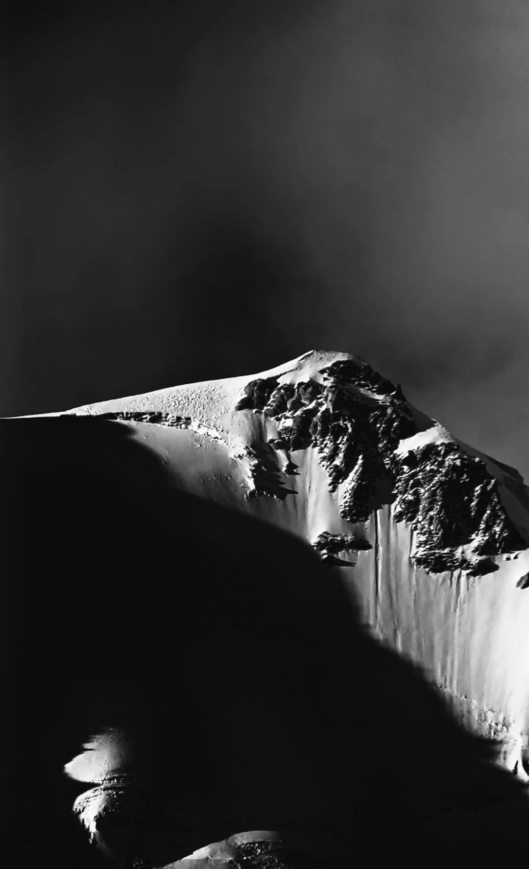 A black and white photograph of a snow-covered mountain with rugged cliffs and steep slopes.