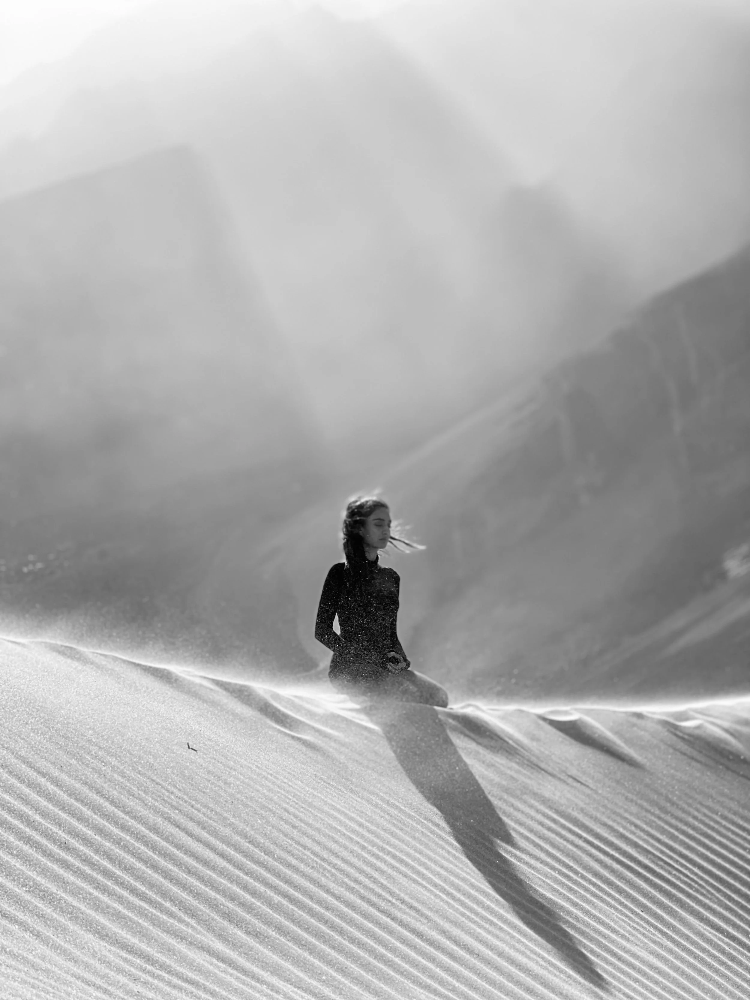 A black and white photograph of a woman on a sandy beach, with her hair blowing in the wind, facing left with her eyes closed.