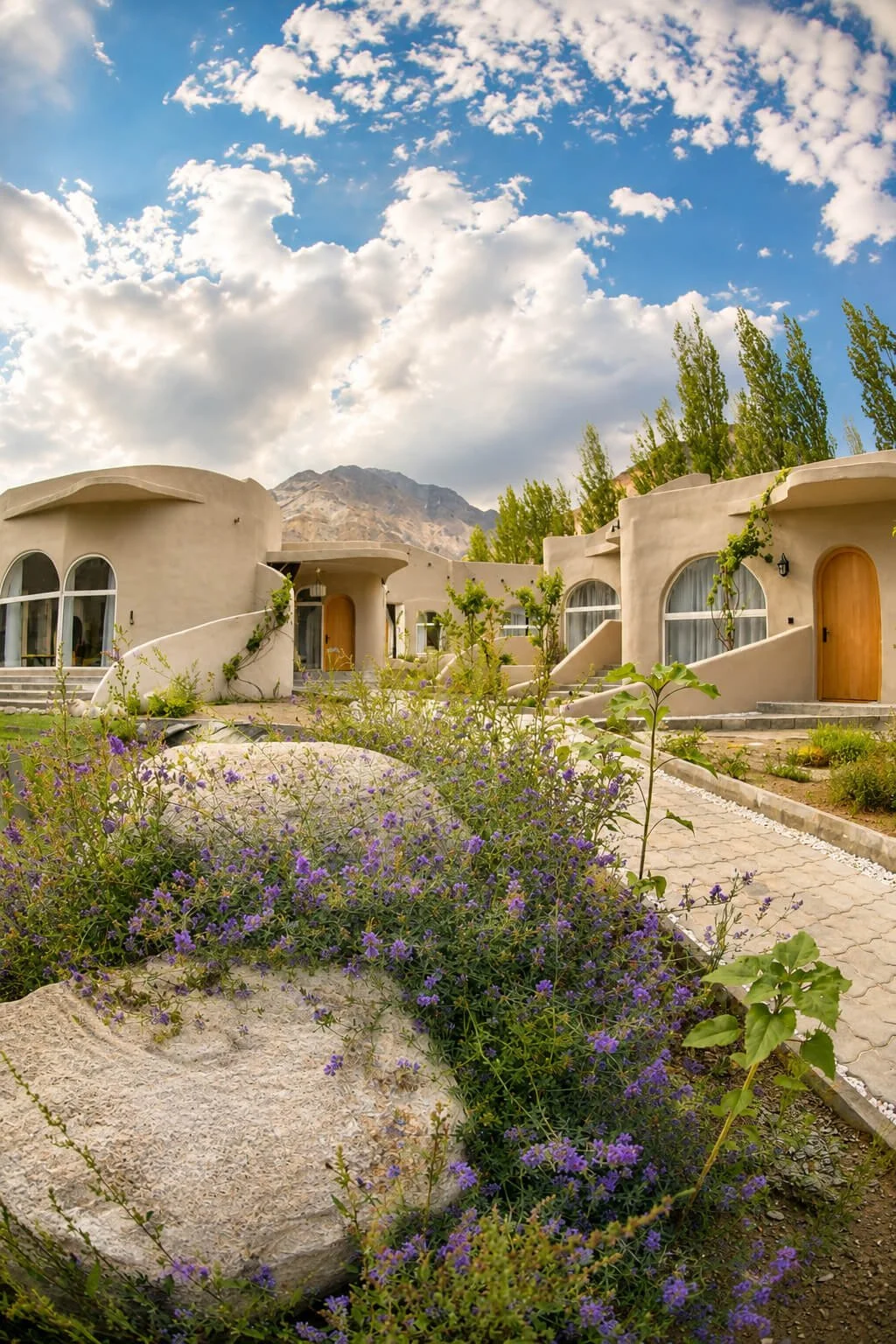 A row of adobe-style houses with rounded walls and arched windows under a partly cloudy sky. Vegetation and purple flowers grow in the foreground.