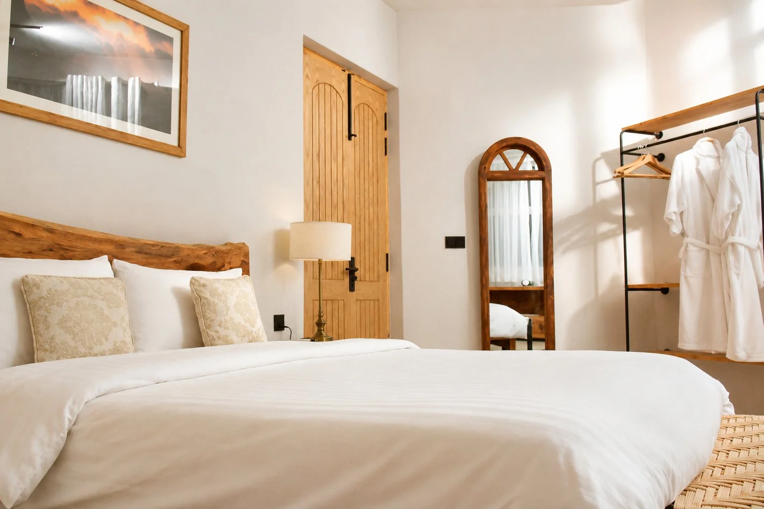 A cozy bedroom with a large bed featuring white linens and decorative pillows, a wooden headboard, a bedside lamp, a full-length mirror, a robe on a clothing rack, and a framed artwork on the wall.
