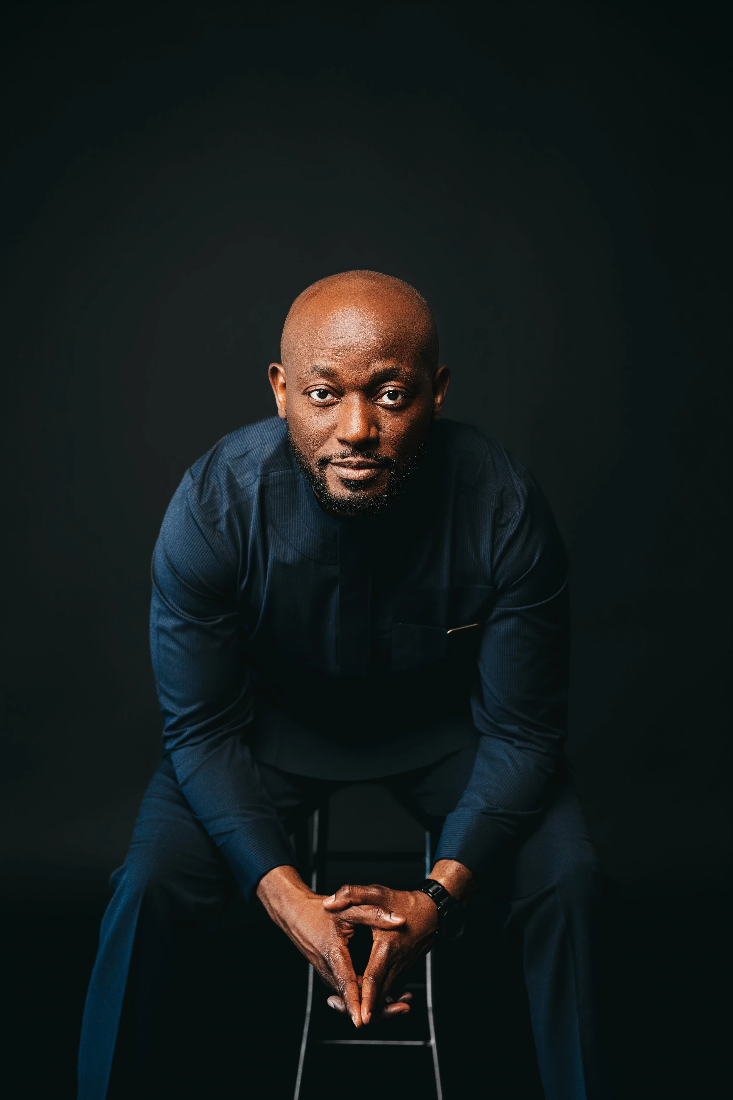 A man with a shaved head and beard sits on a stool, leaning forward with hands clasped. He wears a dark blue shirt and looks directly at the camera against a black background.