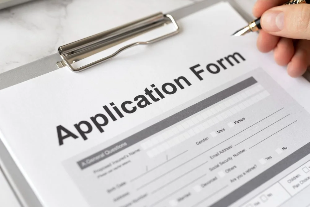 Application form on clipboard with a person's hand holding a pen poised to write.
