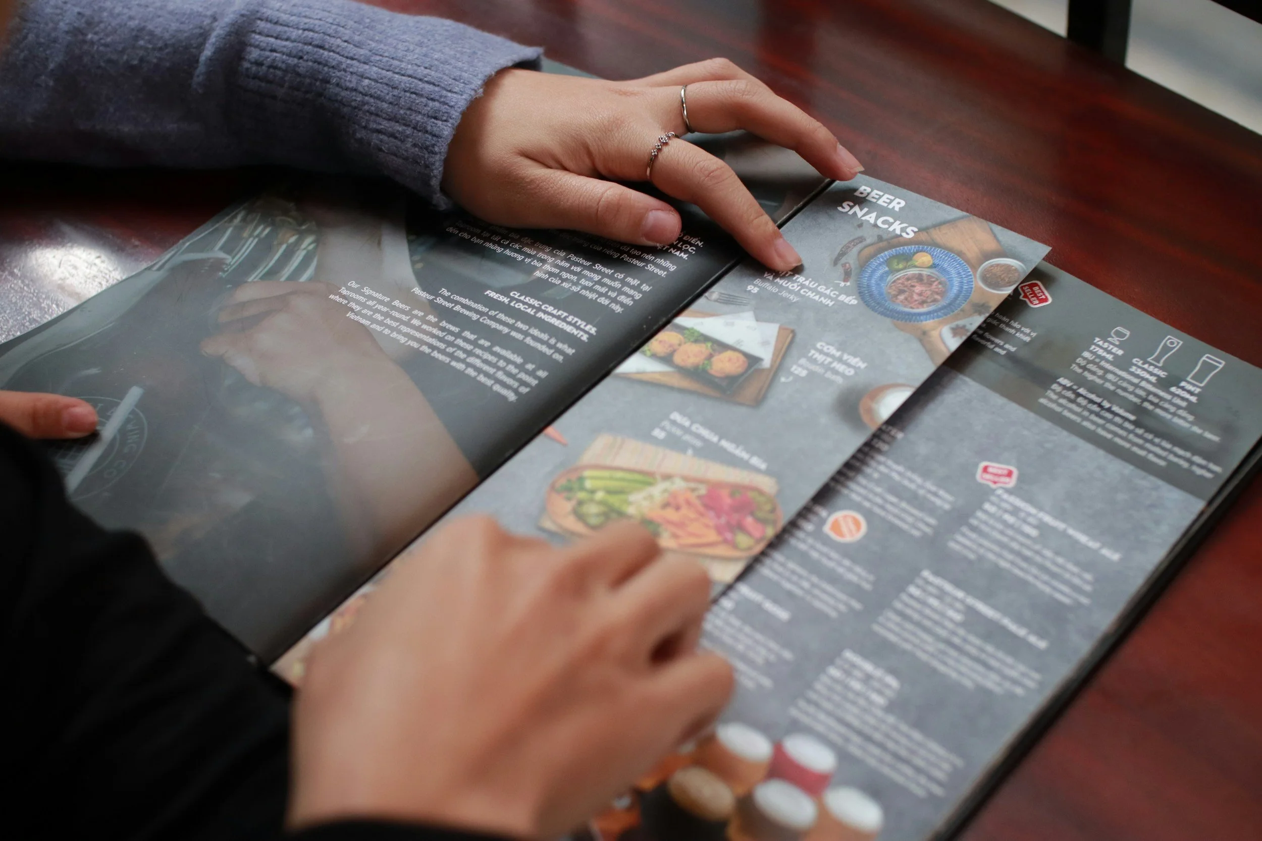 A person with a gray sweater pointing at a menu on a dark wooden table, with the menu showcasing drink options and food images.