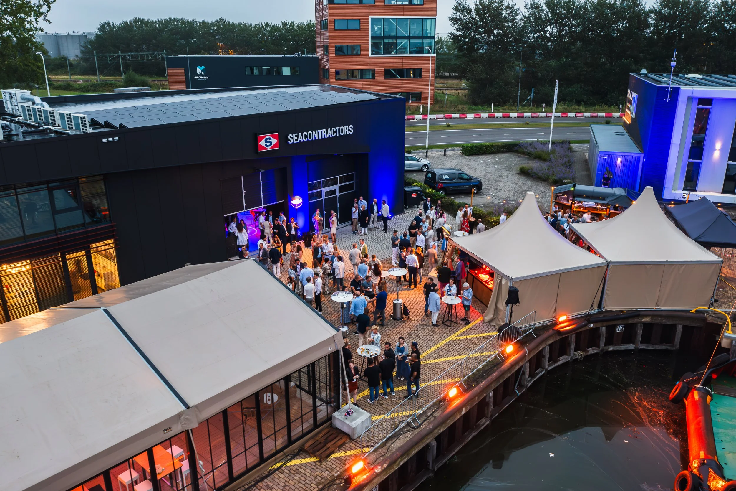 People attending an outdoor event or party with tents, sitting and standing under lit tents against a building with a sign reading 'SeaContracts.' There is a canal with a boat and a roller coaster track in the foreground, and modern buildings and greenery in the background.
