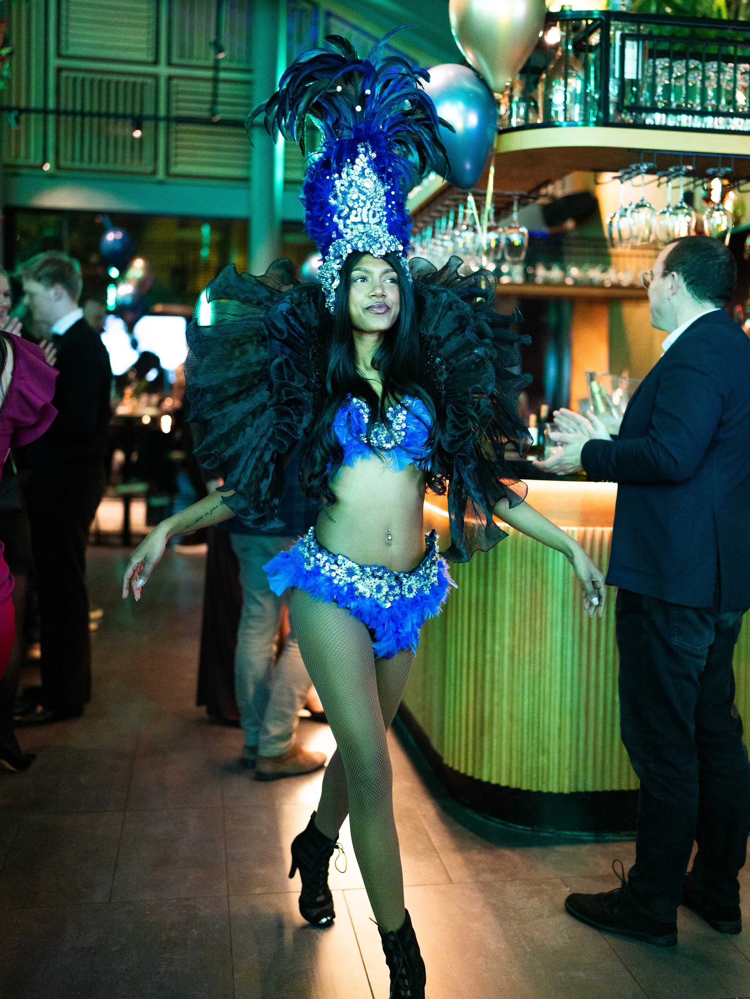 A woman dressed in a vibrant, elaborate costume featuring a large feathered headdress, sequined top, ruffled skirt, fishnet stockings, and combat boots, performing in a lively indoor setting surrounded by balloons and other party-goers.
