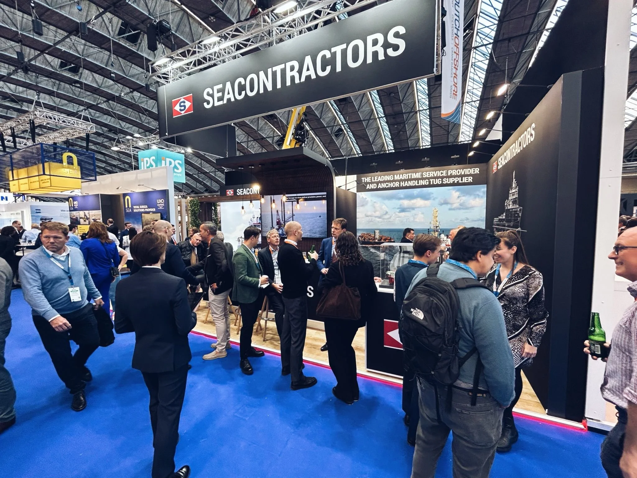 A busy indoor trade show with various booths and many attendees walking and networking. The prominent booth is for SEACONTRACTORS, with a large black sign and ocean-themed graphics on the back panel. The ceiling is industrial with visible metal frameworks and lighting.