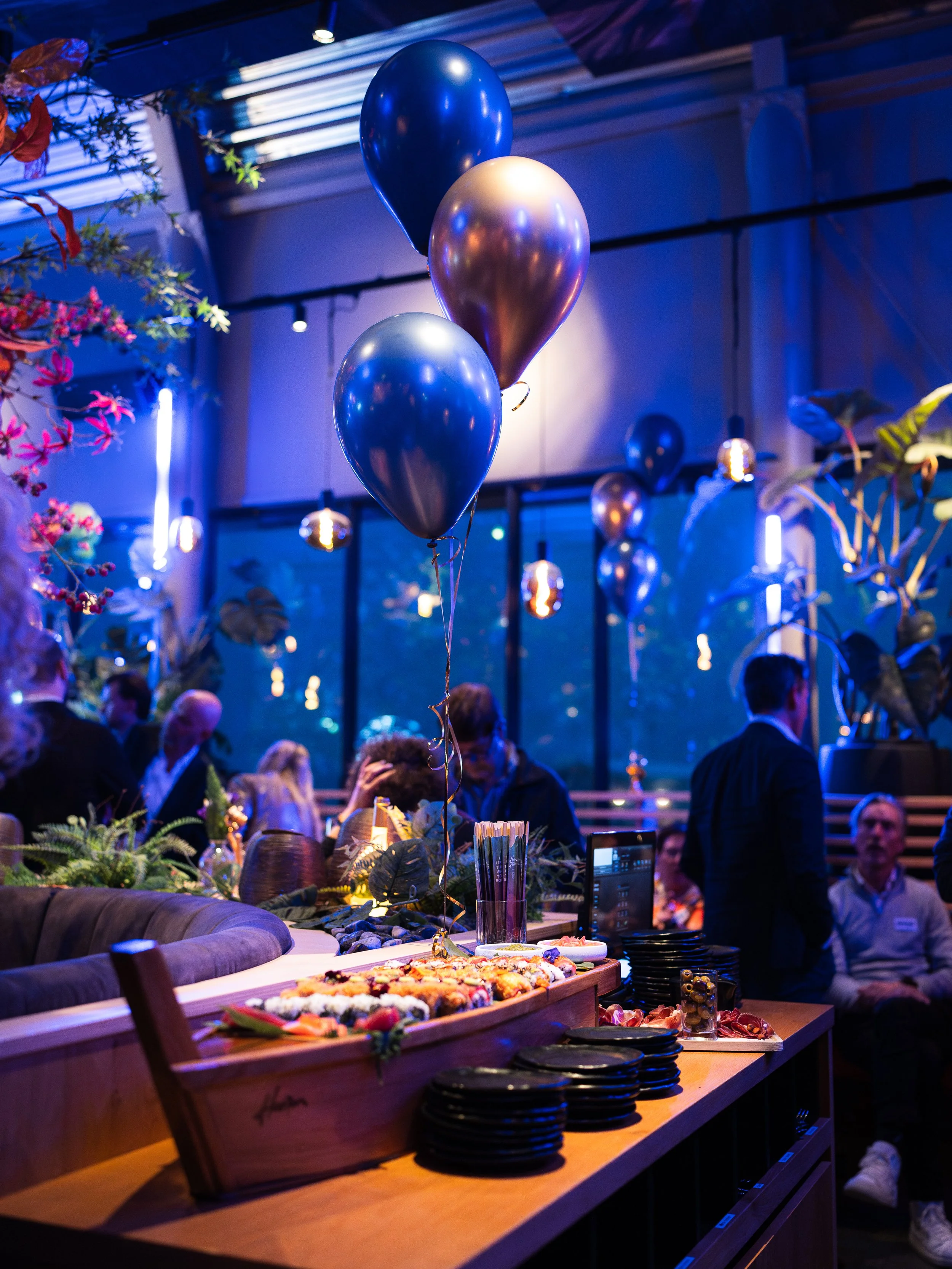 Decorated indoor party scene with blue, gold, and black balloons, people mingling, and a buffet table with food, during evening or nighttime with atmospheric lighting.