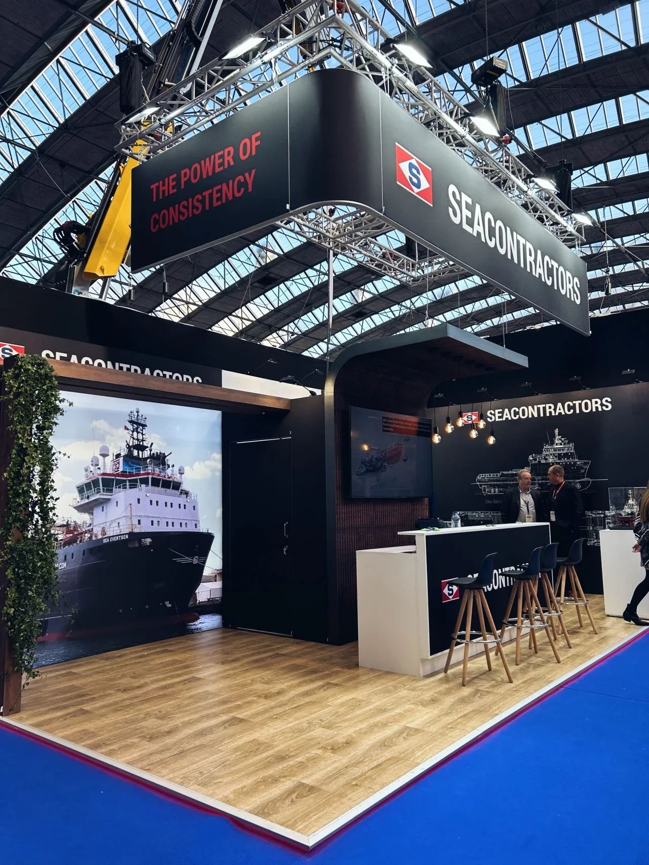 Trade show booth for SEACONTRACTORS with a maritime theme, featuring an image of a ship and a diagram of a ship on the wall, along with seating and informational displays.