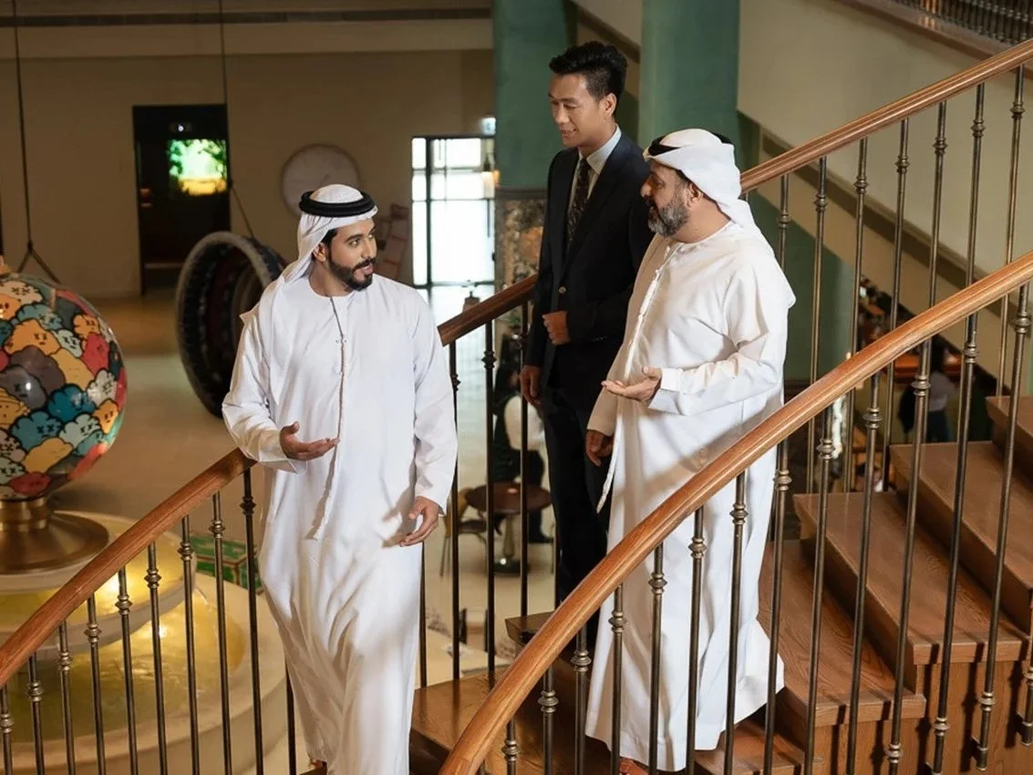 Three men engaged in conversation on a curved staircase: two dressed in traditional Middle Eastern attire and one in a business suit, inside a decoratively decorated building.