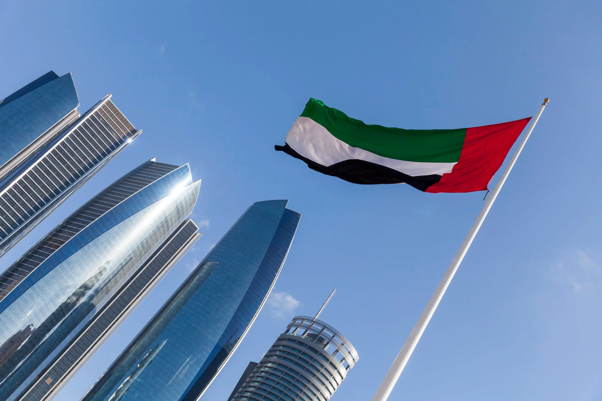 A United Arab Emirates flag flying in front of modern skyscrapers against a blue sky.