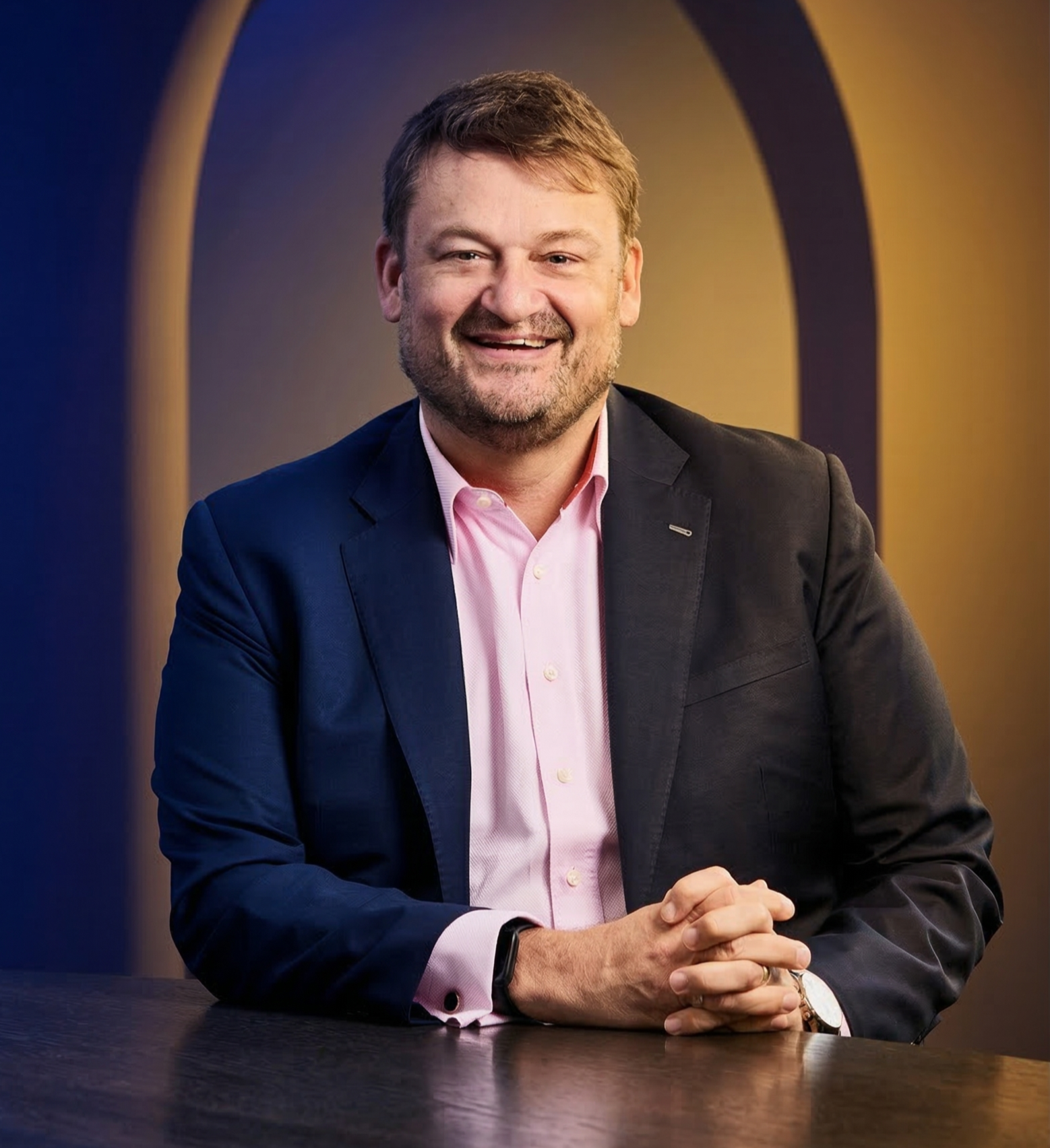 A smiling man in a dark suit and pink shirt sitting at a table with his hands clasped in front of him, in front of a dark background with an illuminated arch.