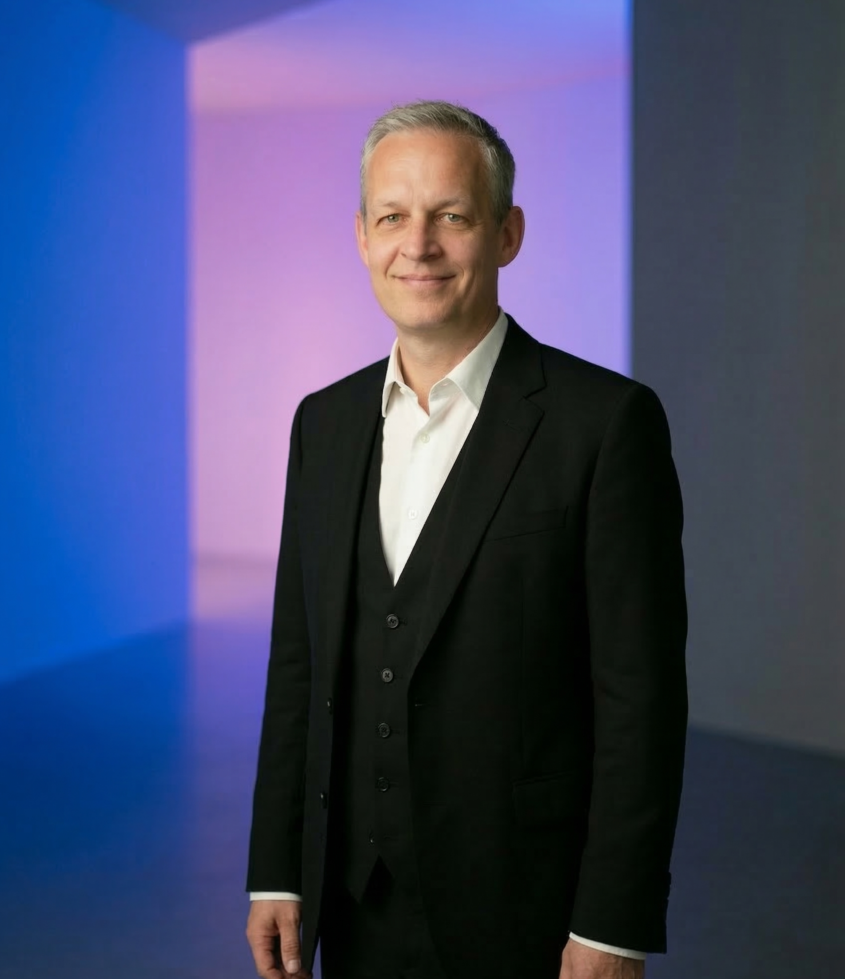 A man in a black suit and white shirt standing in front of a colorful, blurred background.