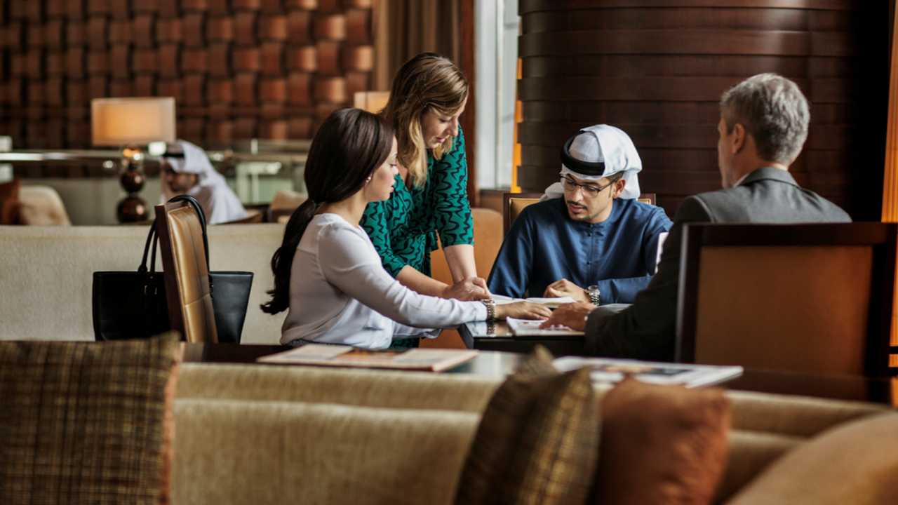 A group of four people seated around a table in a restaurant, engaged in a business discussion. One woman is standing and pointing at documents while others listen and look at the papers.
