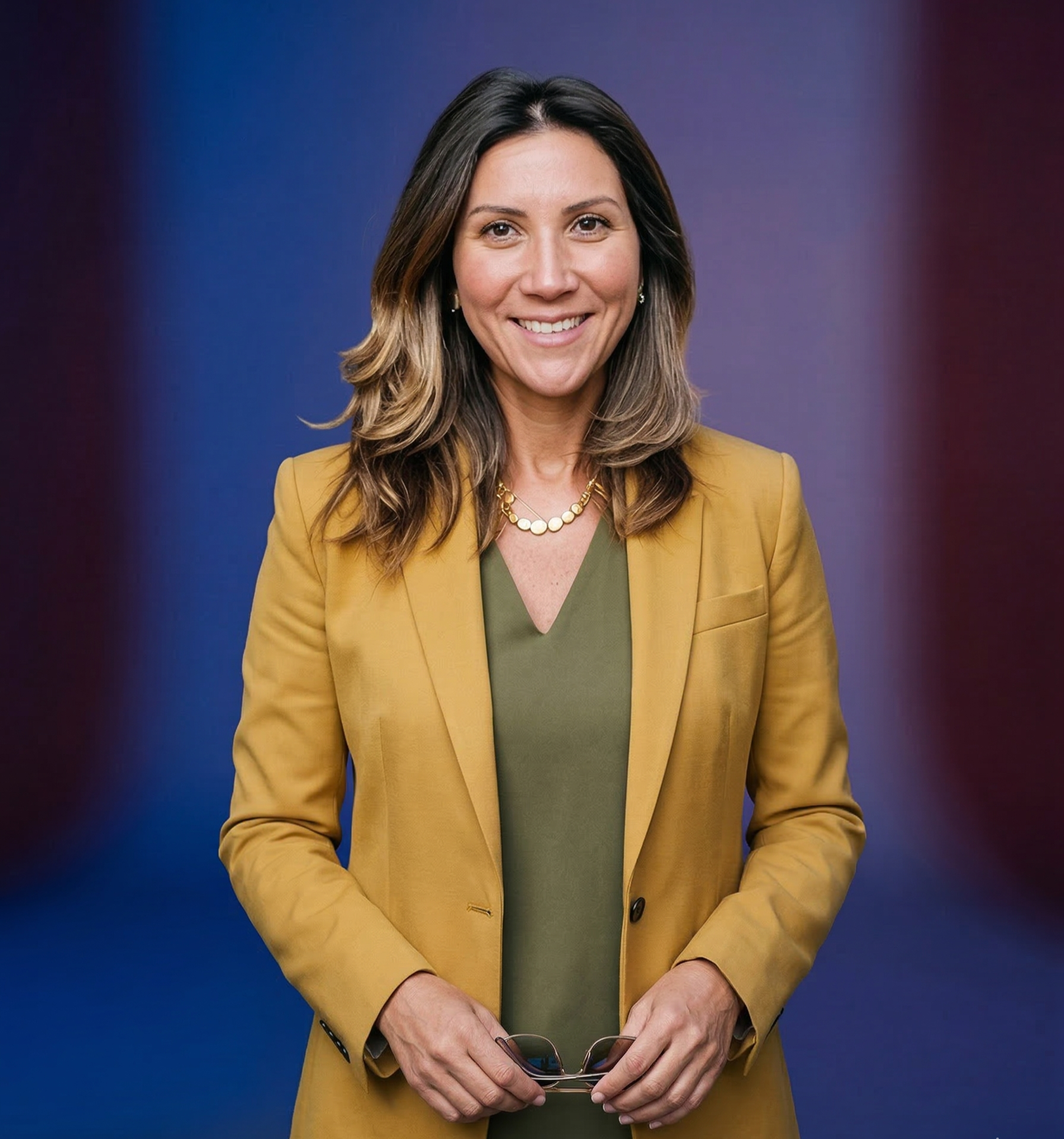 A woman with medium-length brown hair smiling, wearing a mustard yellow blazer over an olive green top, holding glasses, standing against a blurred background with blue and red tones.