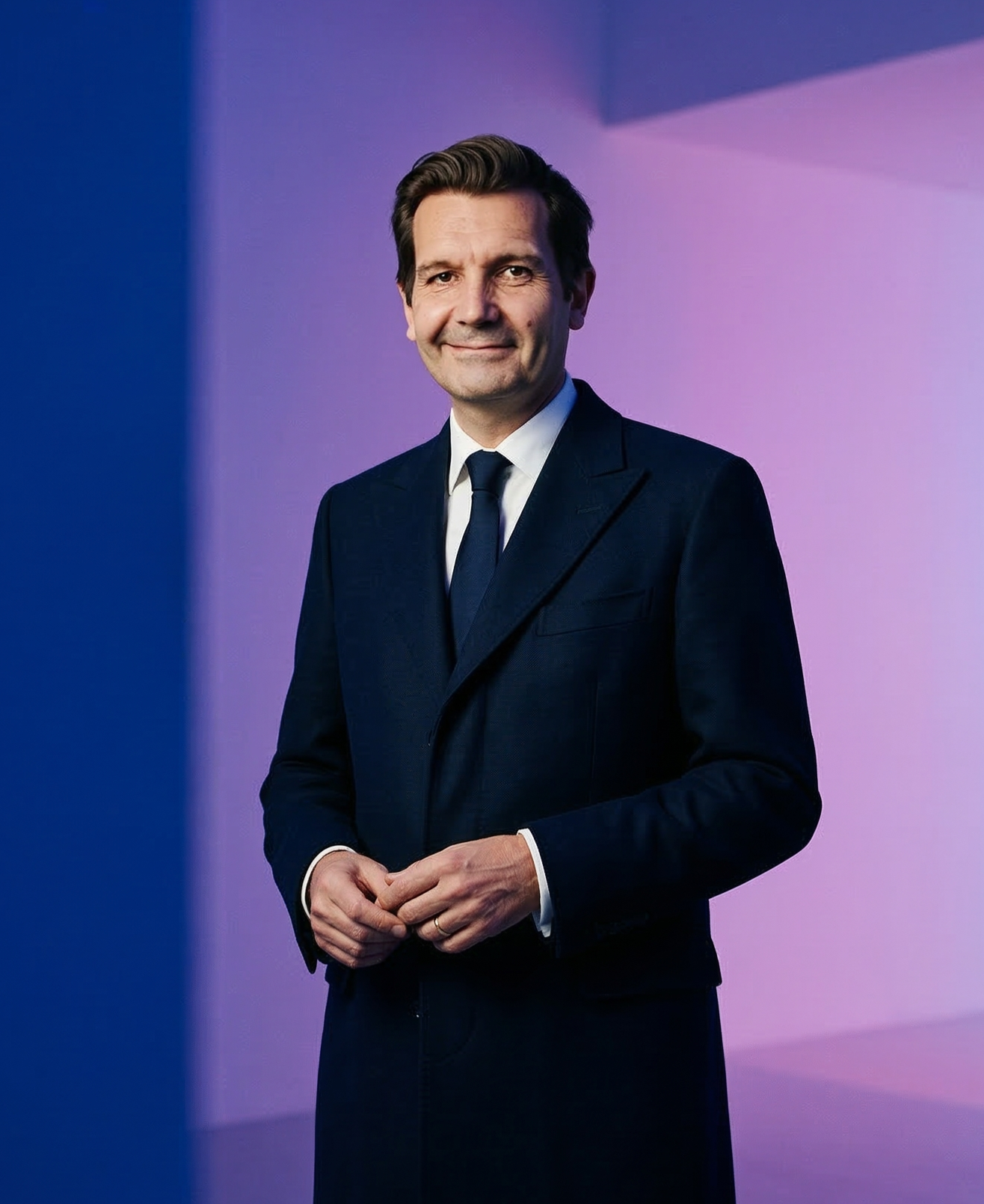 A man in a dark suit with a white shirt and dark tie standing against a gradient background of purple and blue. He is smiling and looking at the camera.