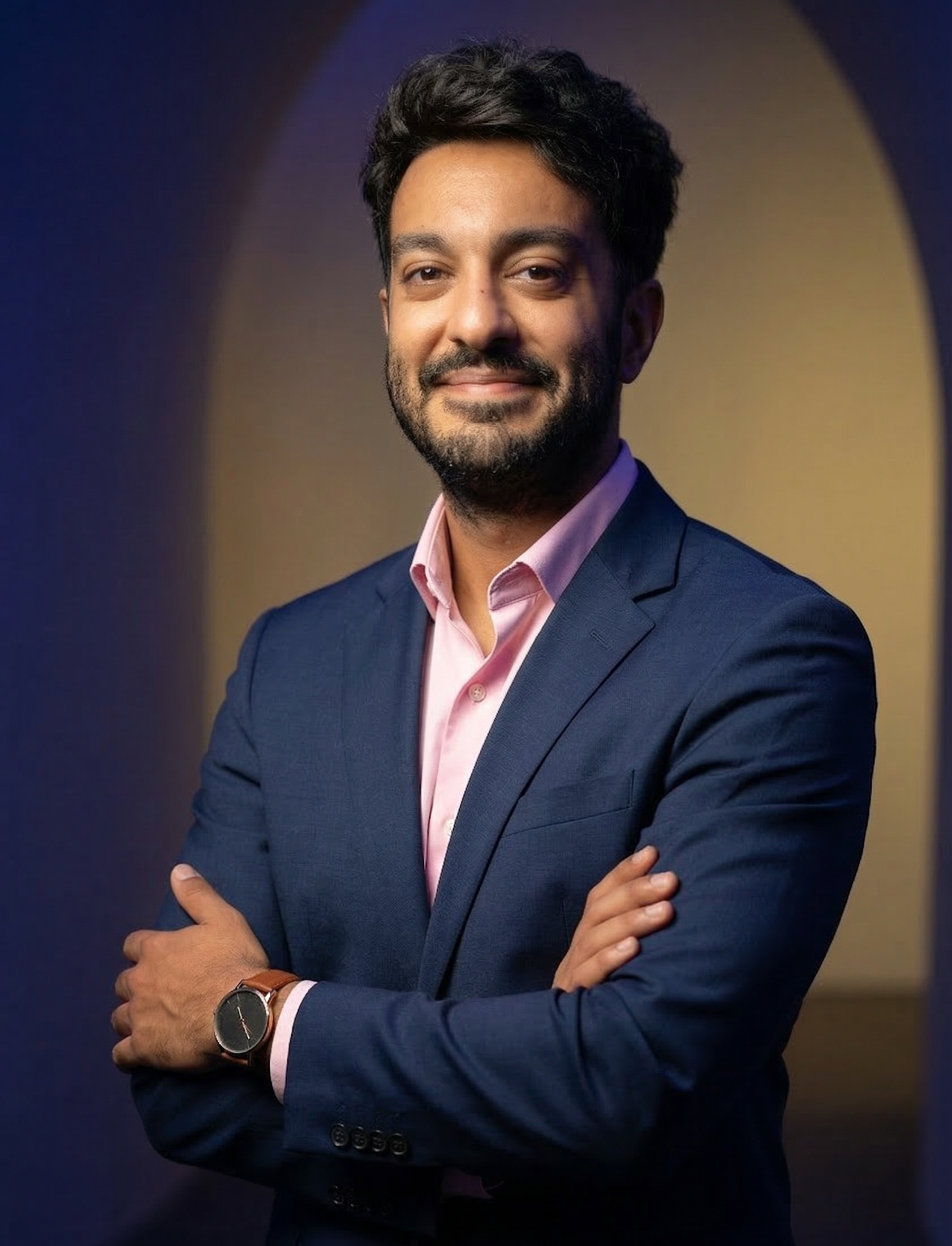 A man in a navy suit and pink shirt with crossed arms, smiling and looking at the camera, standing in front of a colorful background with blue and yellow hues.