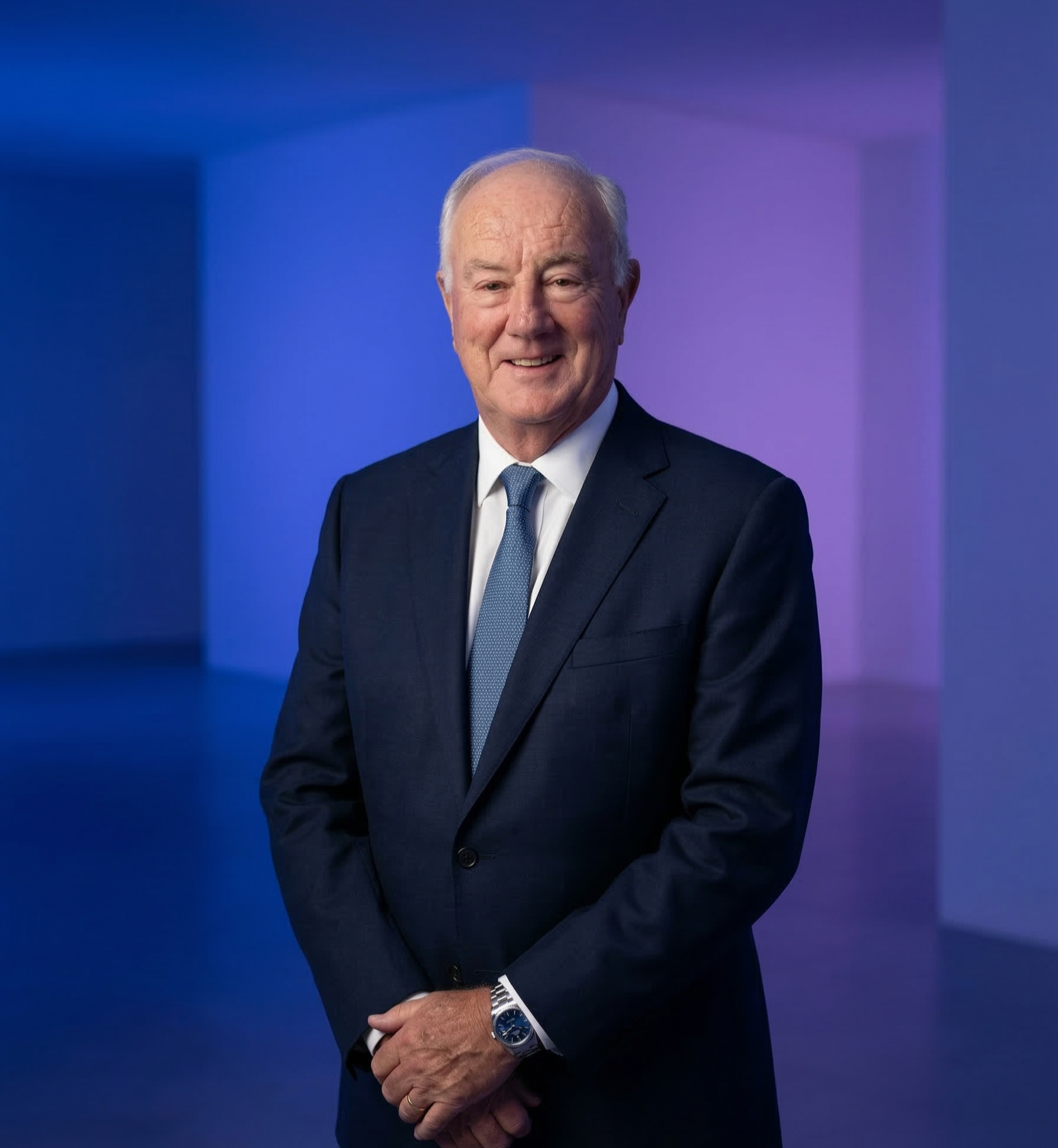 A smiling elderly man in a dark suit and tie standing in front of a blue and purple background.