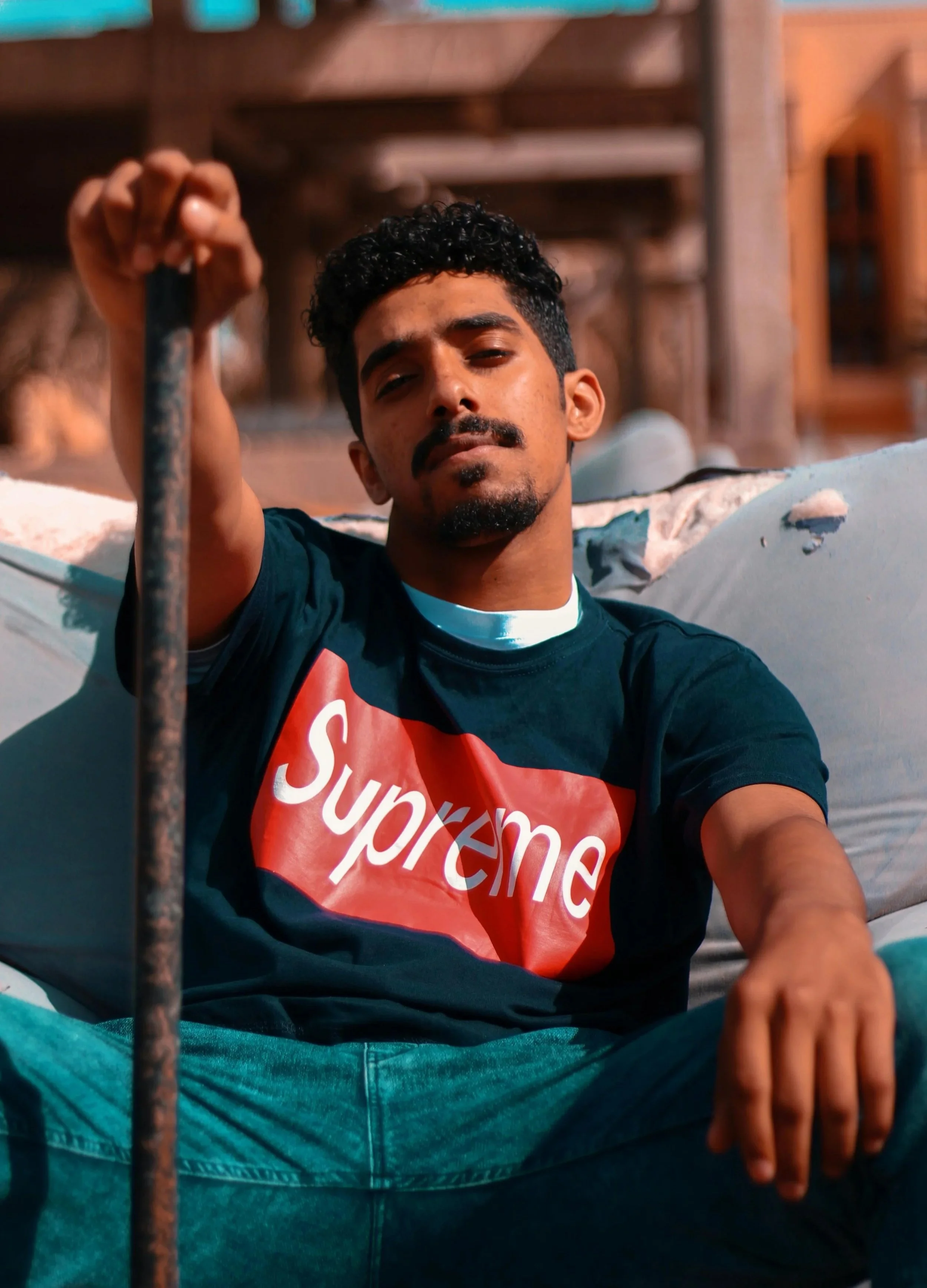A man sitting cross-legged, wearing a black Supreme T-shirt, in a relaxed pose with an outdoor background featuring wooden structures.
