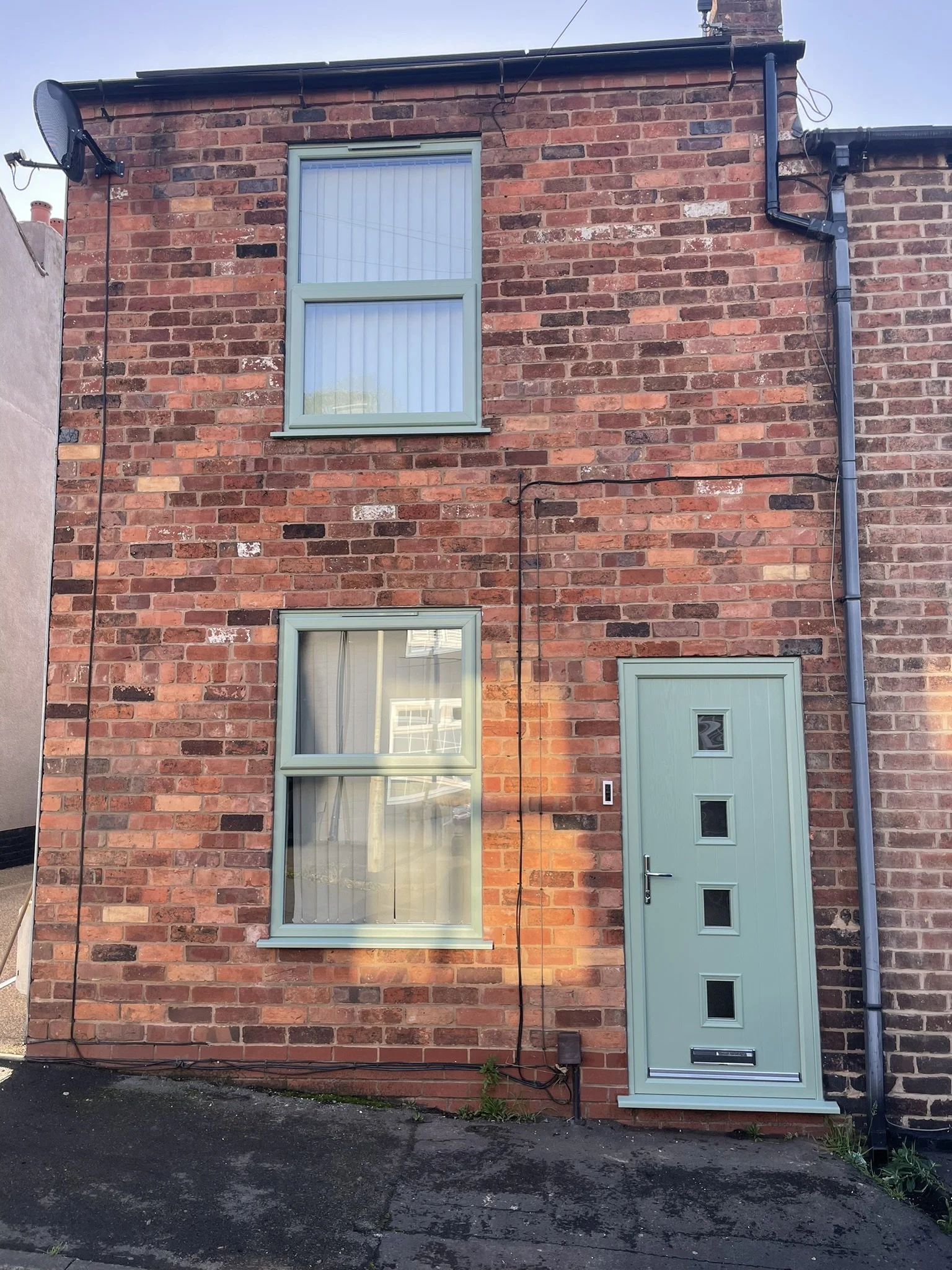 A two-story red brick house with light green window frames and a matching front door. The house has two windows on each floor, a satellite dish on the upper left, and black drain pipes on the right side. The ground is paved with asphalt.