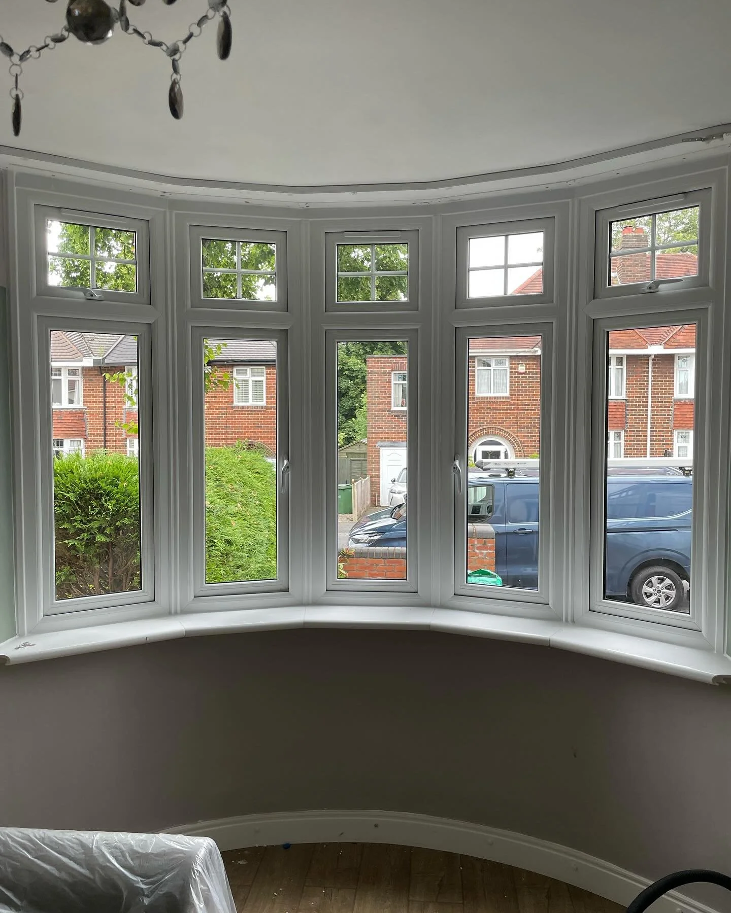 A view from inside a room looking out through a large bay window with multiple panes, showing a suburban street with brick houses, parked cars, greenery, and a driveway.