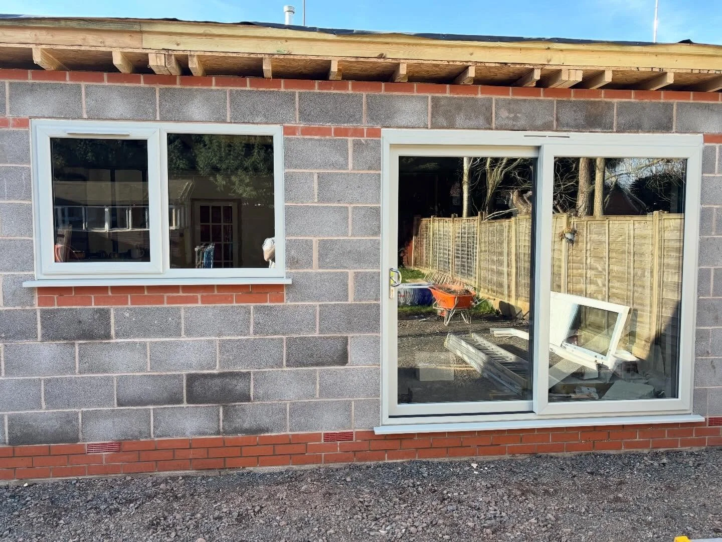 Construction site of a house with an unfinished gray cinder block wall, a double-hung window, and a sliding glass door, with construction materials and tools inside and outside.