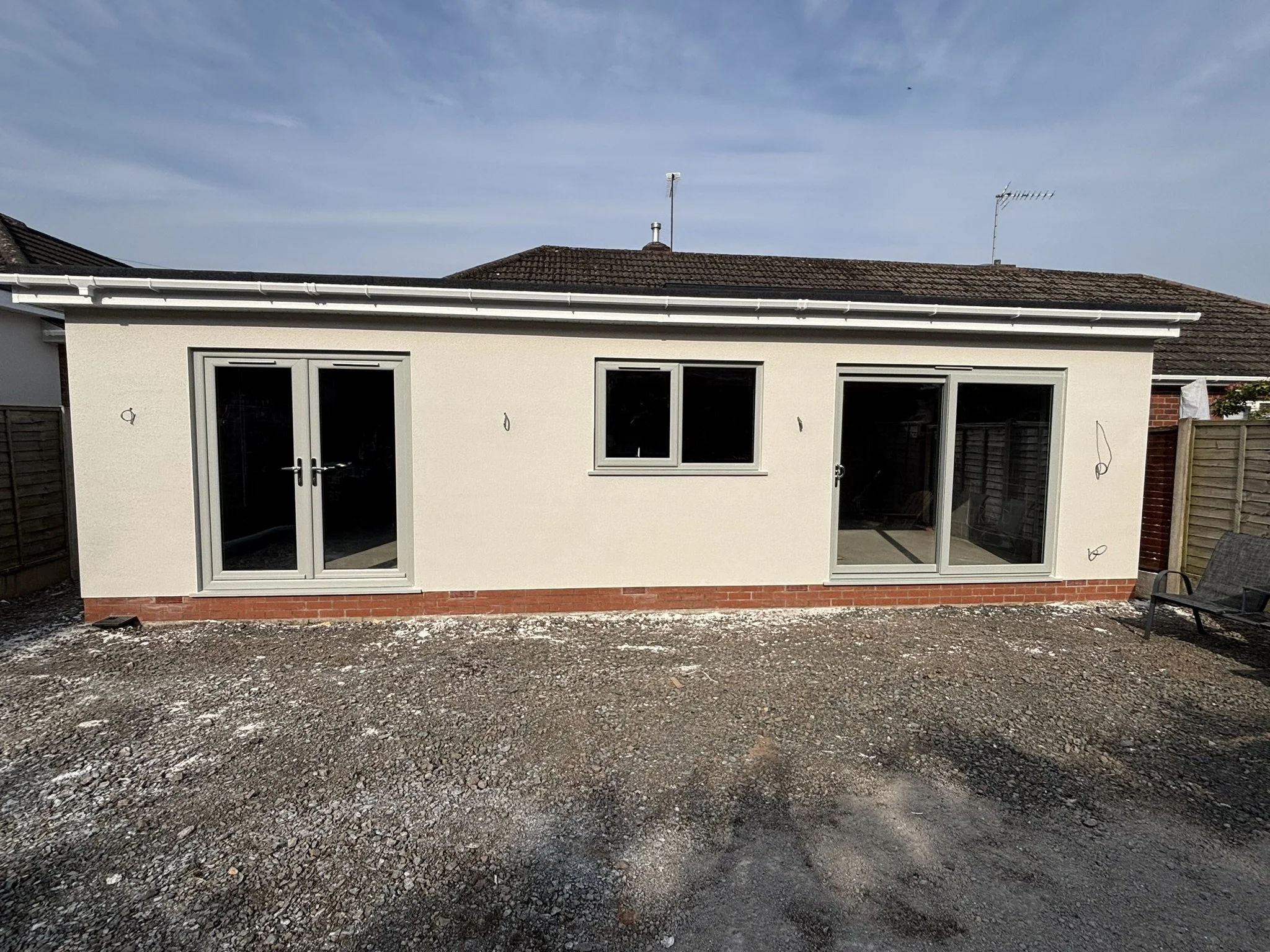 Newly constructed house with white walls, large glass doors, and windows, situated on a gravel surface.