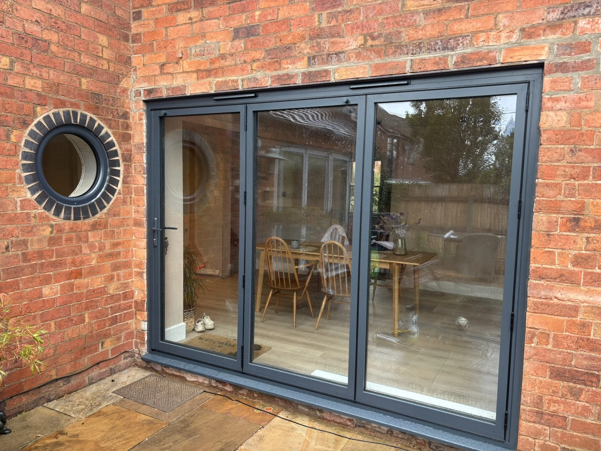 A brick house rear with large sliding glass door leading into a dining room with wooden table and chairs, and a small round window on the brick wall.