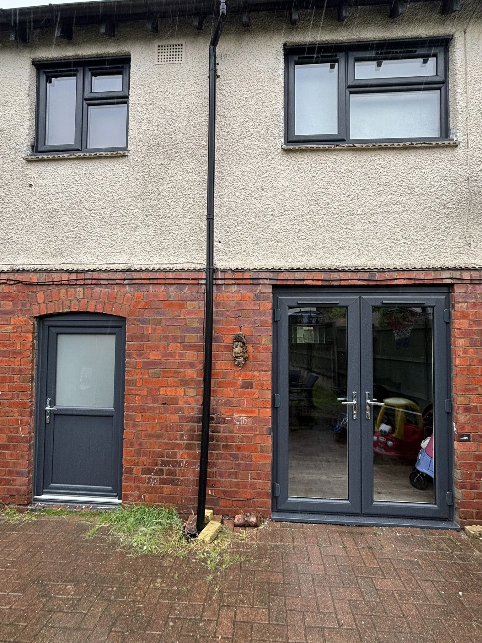 The lower part of a beige and red brick house with two dark blue doors, one normal door on the left and one glass double door on the right, and two windows above. There is a black gutter pipe running vertically between the doors, and small patches of