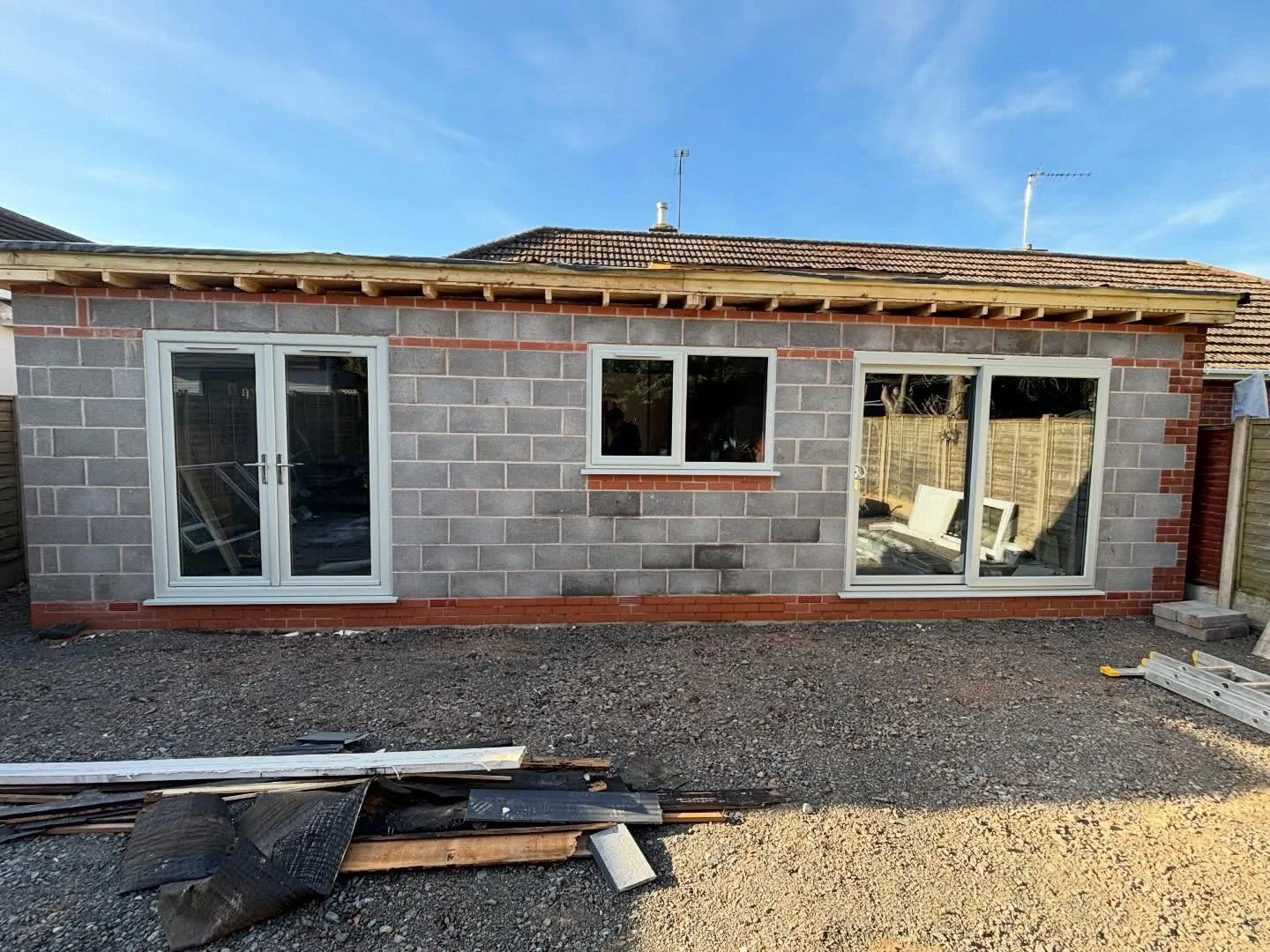Backyard of a house under construction with unfinished walls, two sliding glass doors, one window, construction materials on the ground, and a clear blue sky.