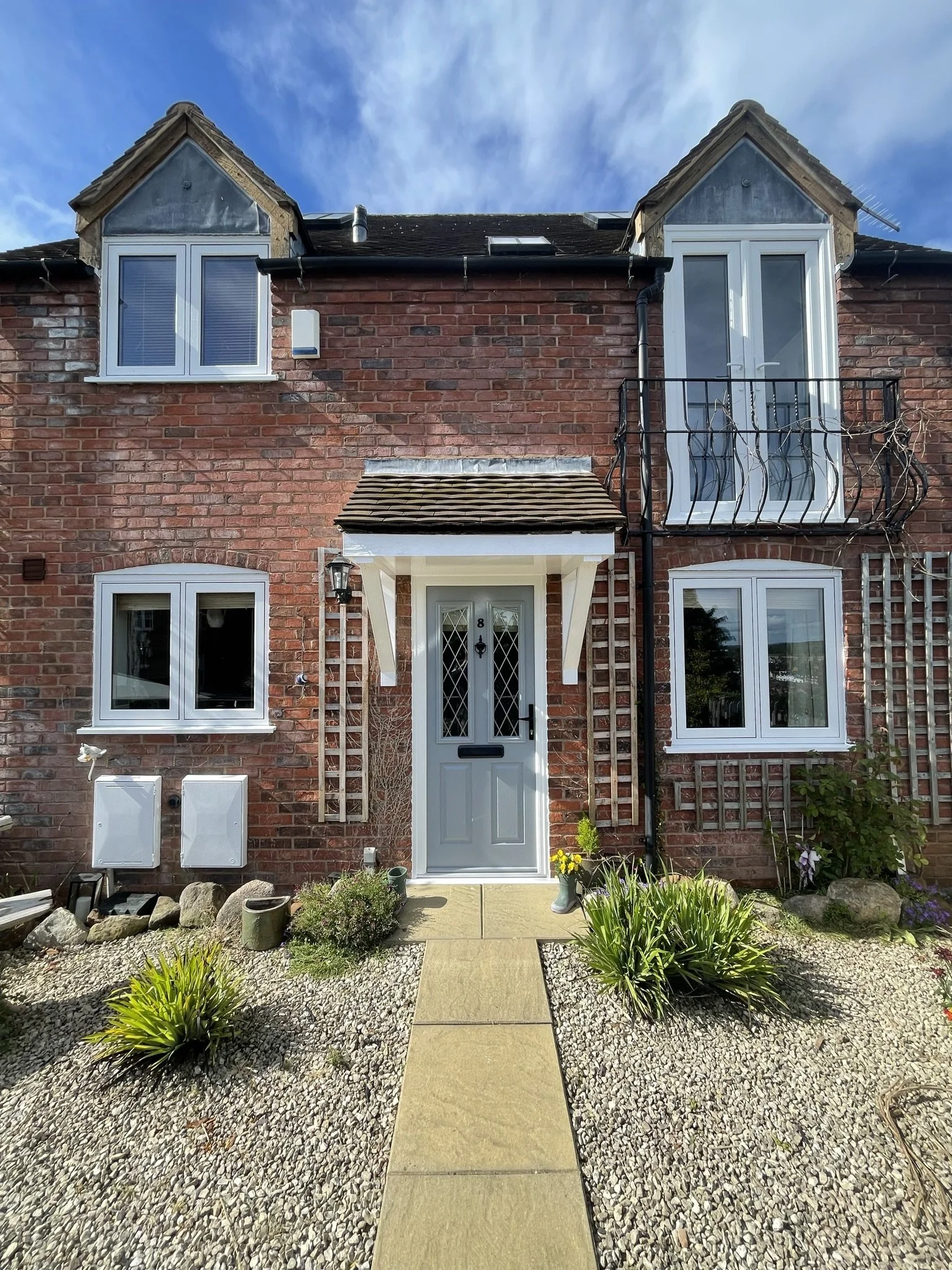 Front view of a brick house with a central white door, three windows, and small upper windows with dormer roofs. The house features a small porch with a brown tiled roof, a black lantern, and a gravel garden with green plants and flowers.
