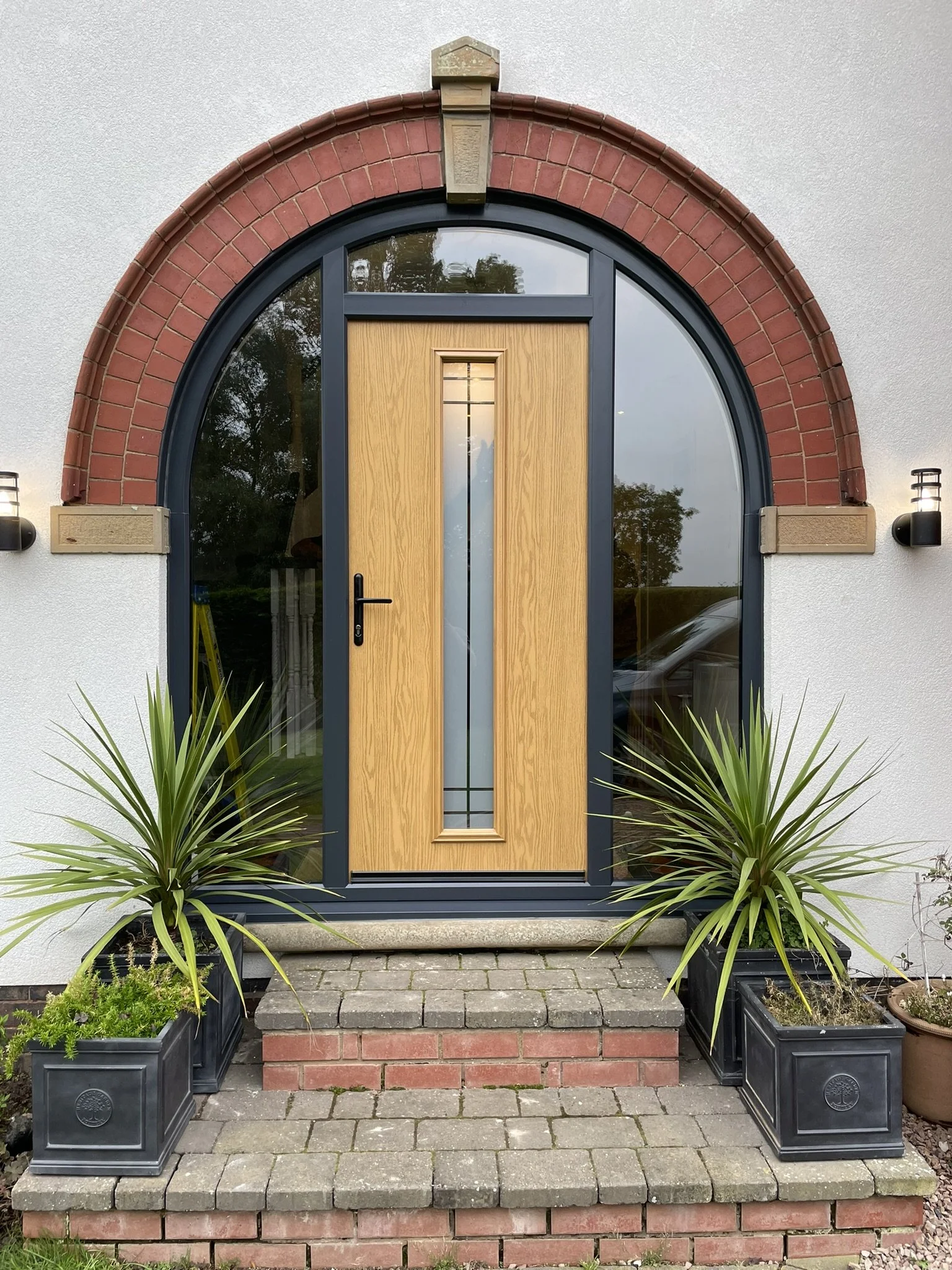 Front entrance of a house with a wooden door, arched window above, brick framing, and surrounding potted plants on brick and stone steps.
