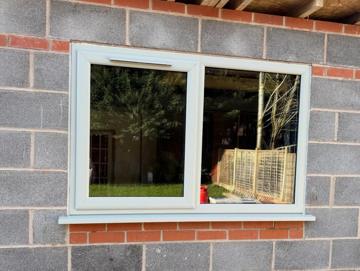 A window set into a gray concrete wall with brick accents, showing a backyard with grass, trees, a wooden fence, and outdoor objects.