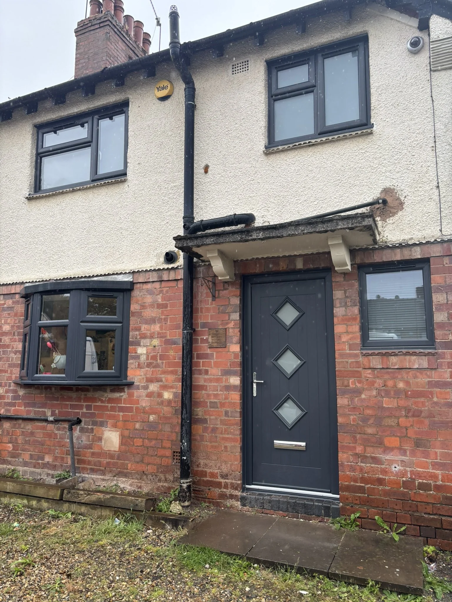 The exterior of a two-story house with a brick and stucco facade, featuring a black front door with diamond-shaped glass panes, three windows, a black drainpipe, and a Yale security alarm box on the wall.