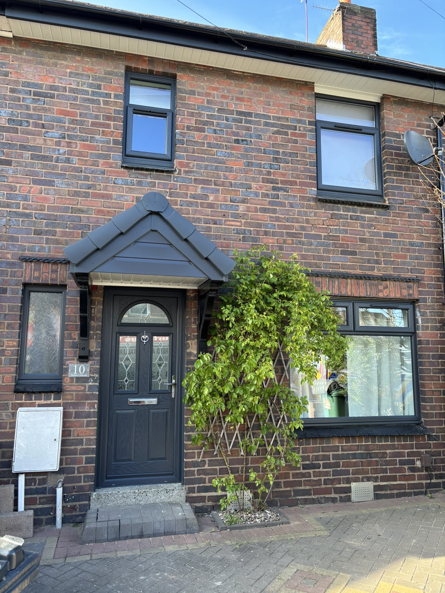 Brick house with black front door, small canopy above, and two upper windows. A green leafy plant grows next to the door. The sidewalk and part of the street are visible in front.