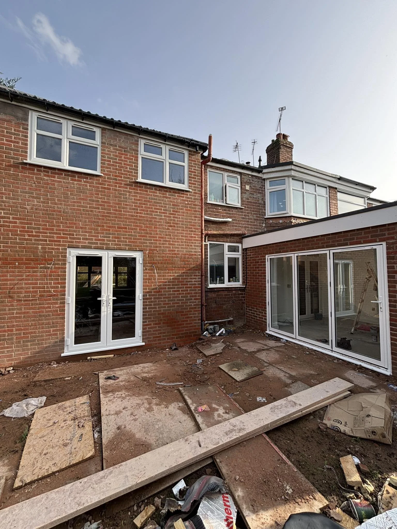 Backyard of a brick house under renovation, with construction materials and dirt on the ground, and large glass doors and windows installed.