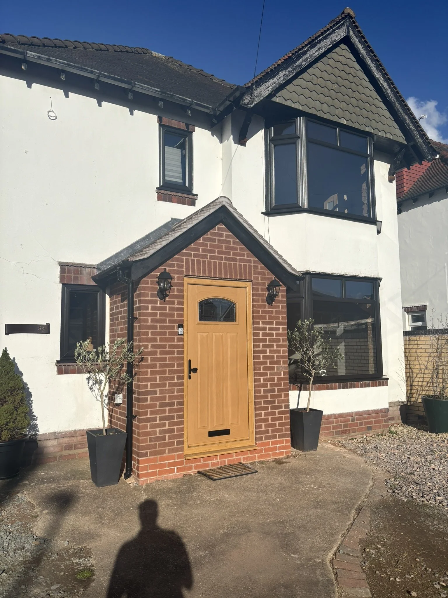 Front view of a two-story house with a brick and white exterior, black window frames, and a wooden front door. There are two potted plants near the doorway, and the house has a sloped roof with dark shingles.